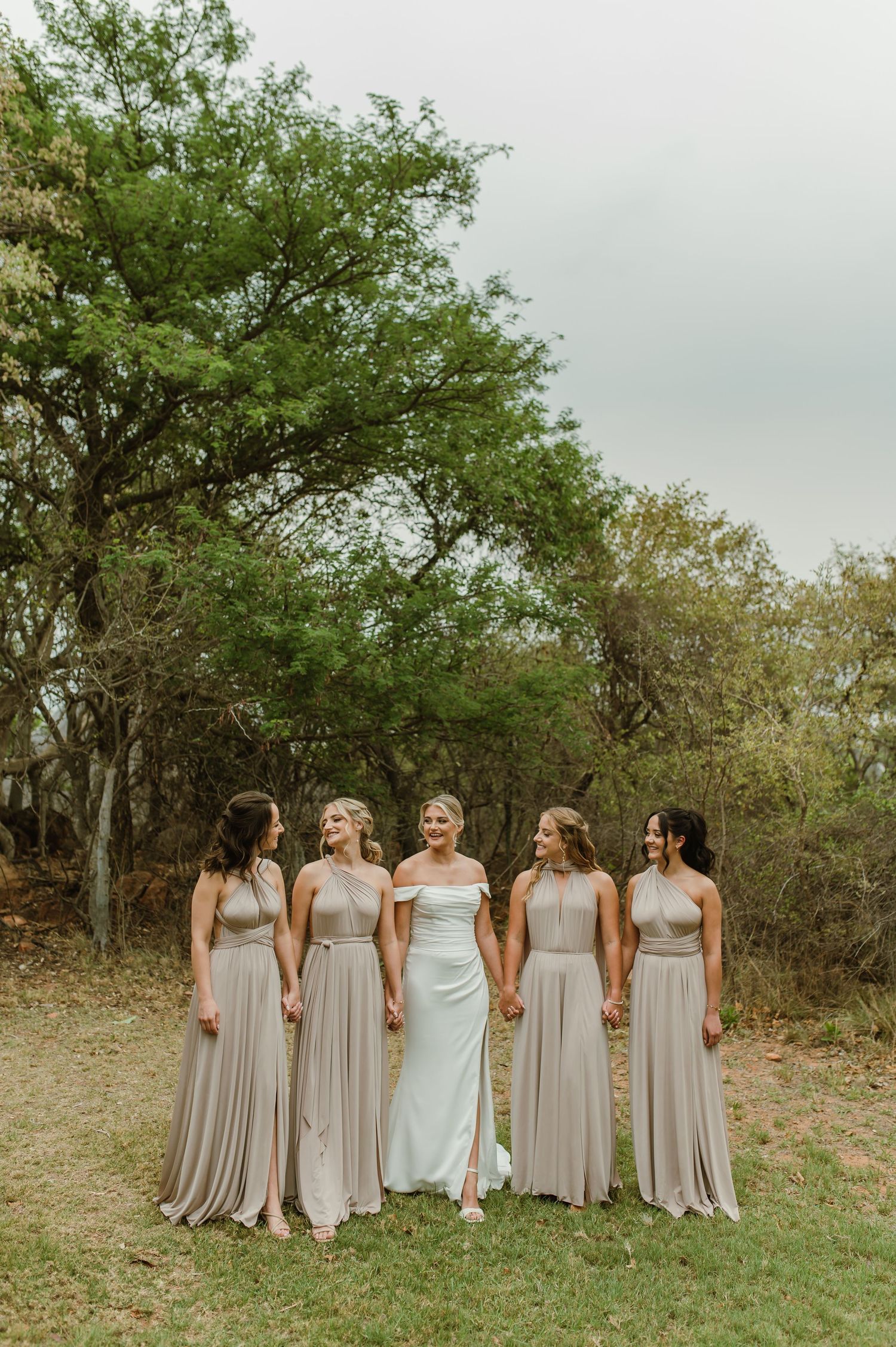 Wedding party poses together in elegant neutral-toned dresses against a natural wooded backdrop.