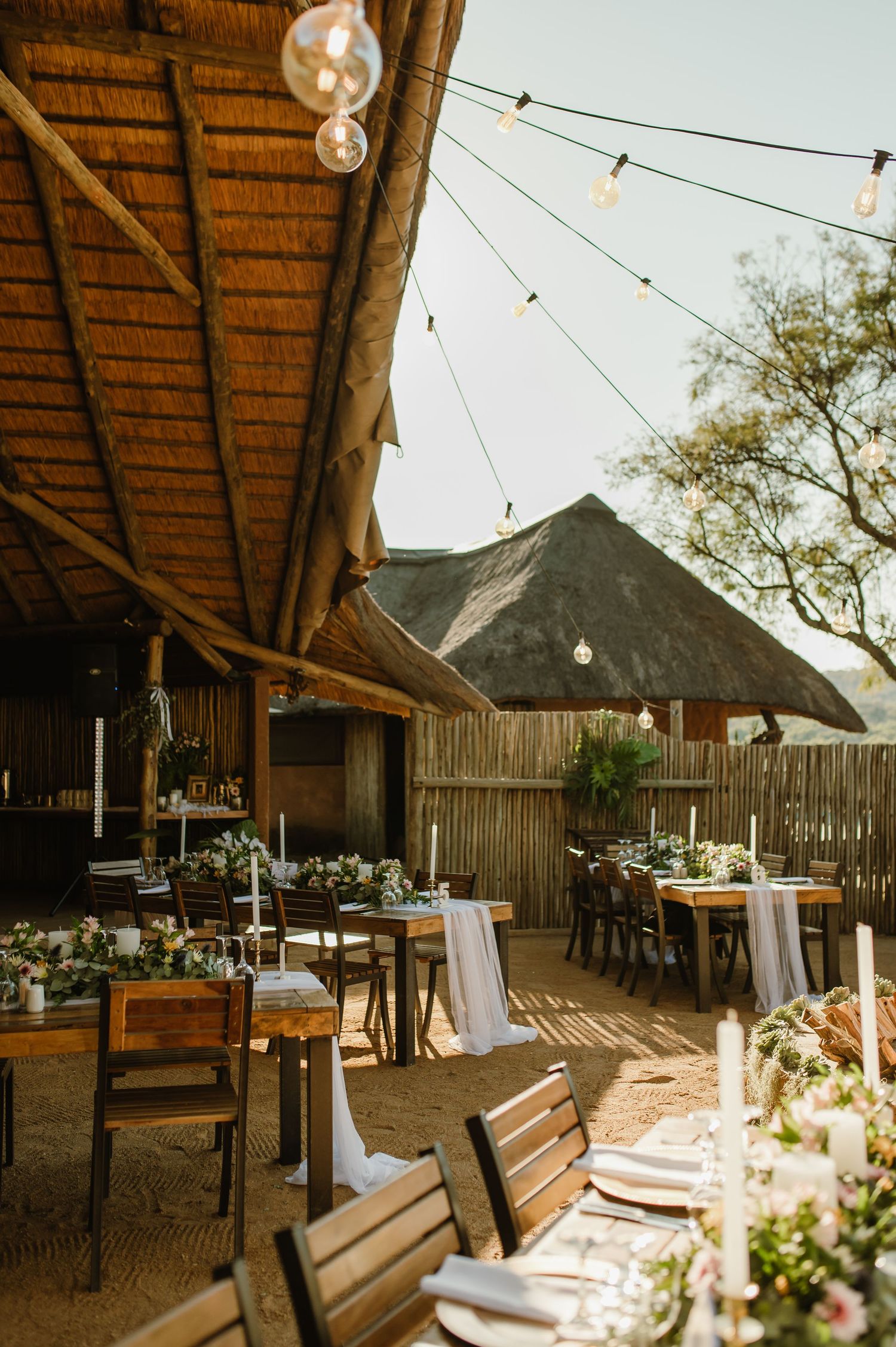 Rustic outdoor wedding venue with thatched roof pavilion and string lights at sunset.