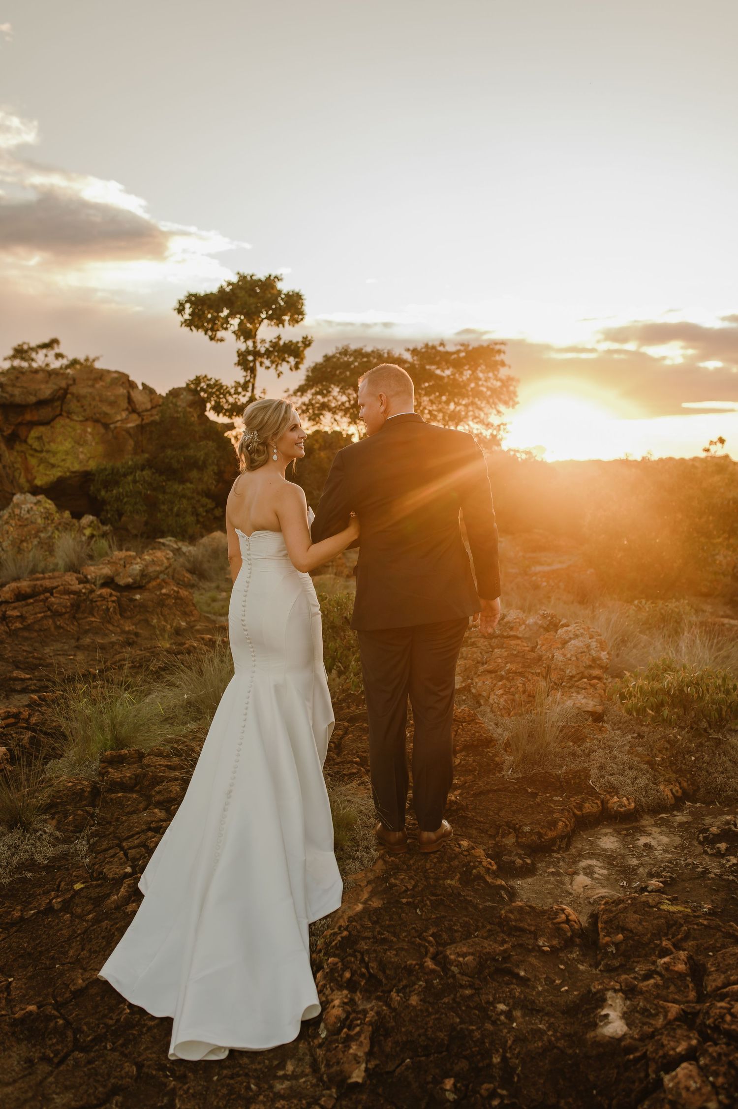 Newlyweds hold hands while walking through rugged terrain during a dramatic golden hour sunset.