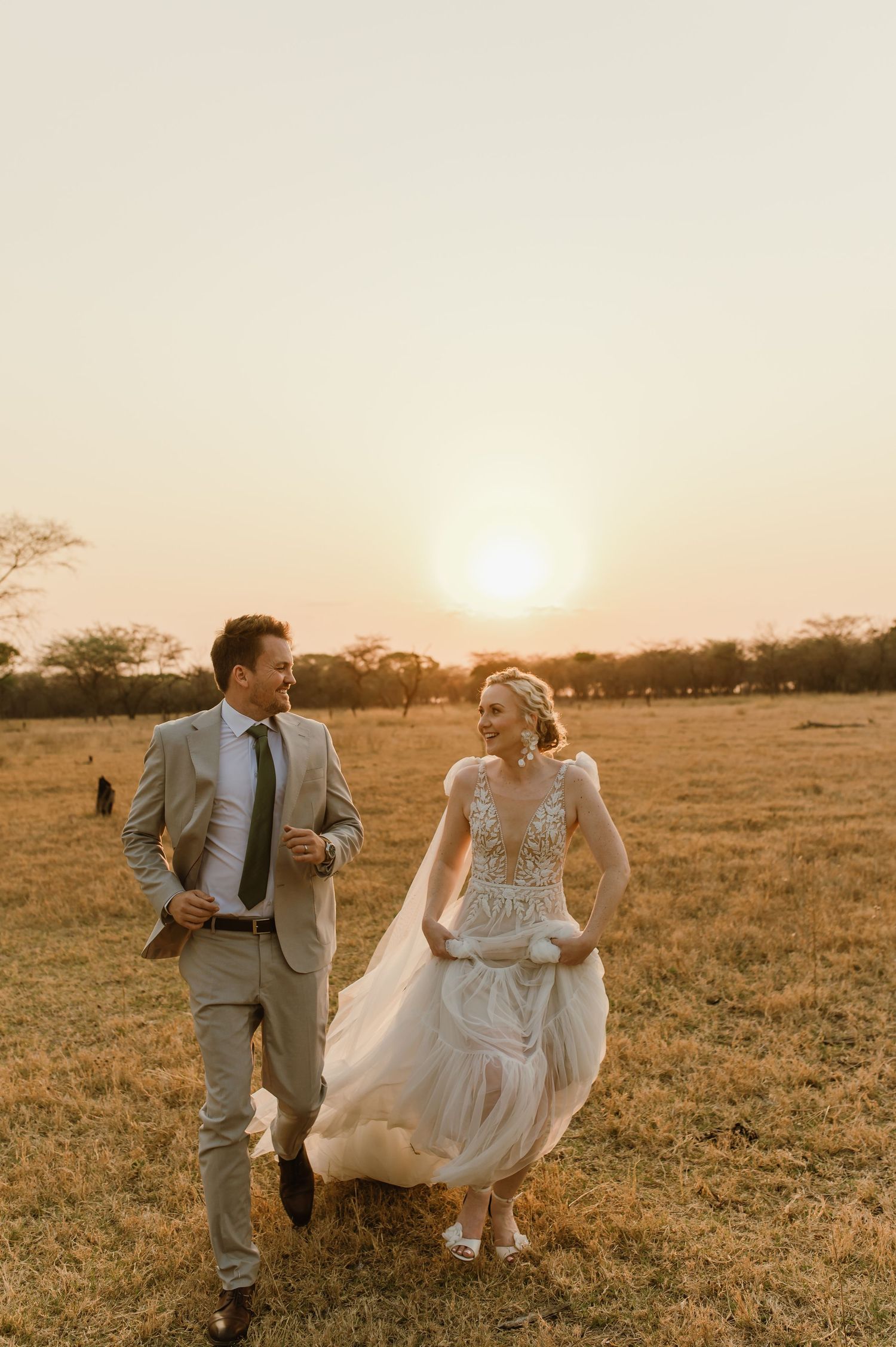 A joyful couple runs through a sun-soaked African savanna field during their wedding celebration.