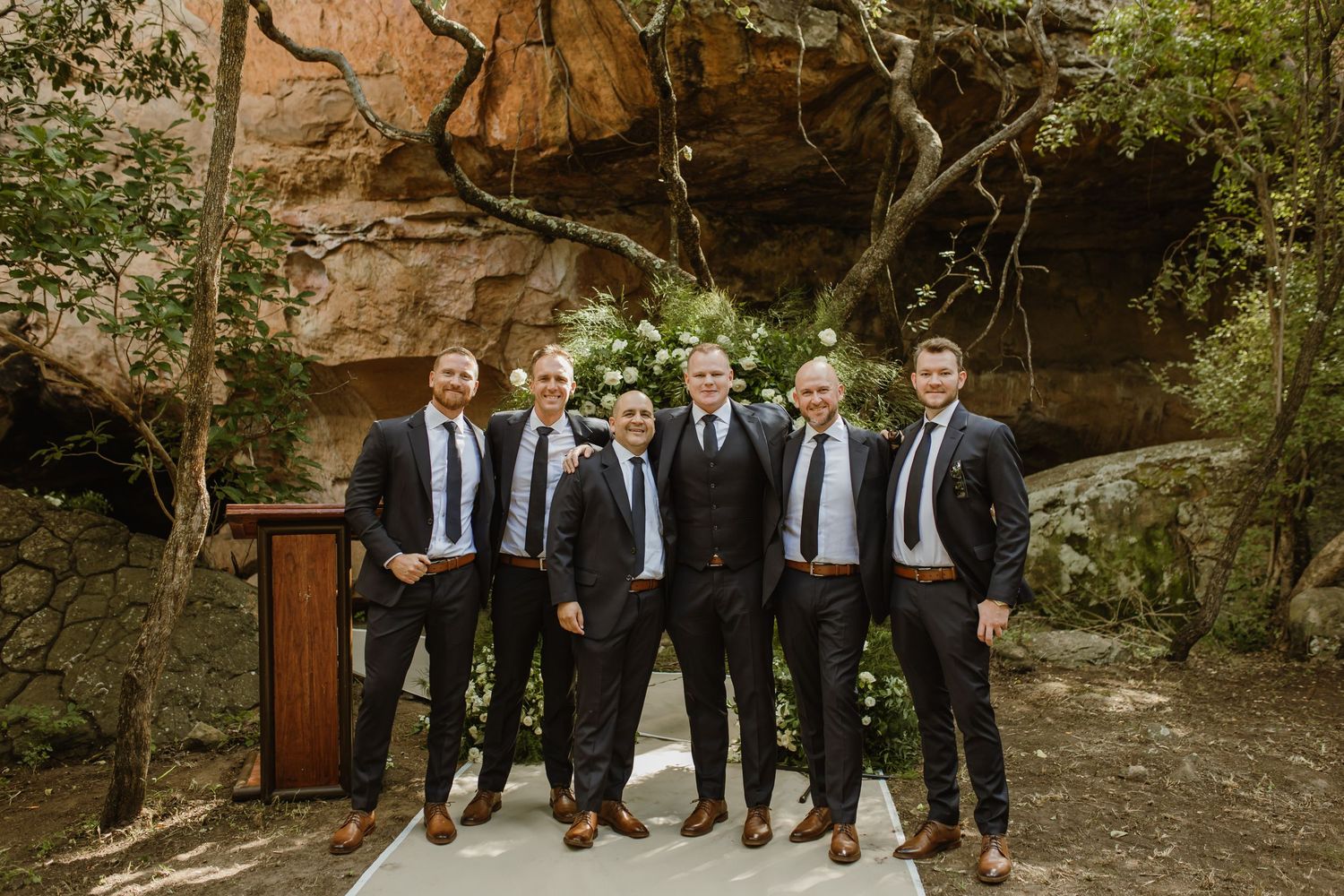 Groomsmen in dark suits pose together against a rocky outdoor backdrop.