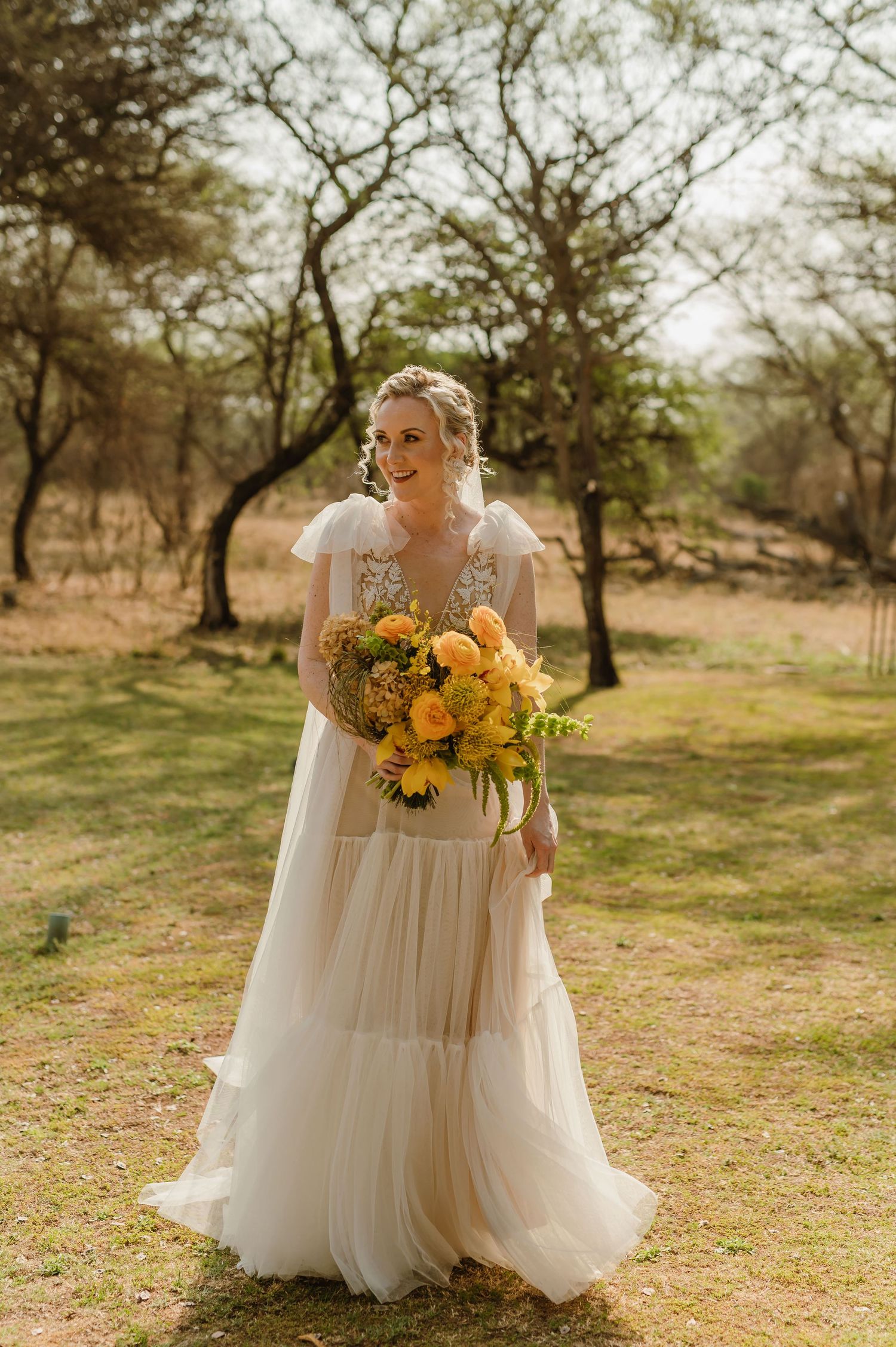 Bride in flowing white dress carries vibrant yellow sunflower bouquet in outdoor setting.