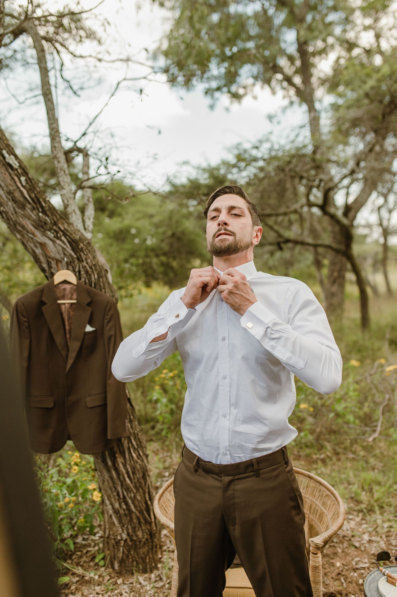 Person adjusting collar of white dress shirt while standing outdoors with suit jacket hanging on tree.