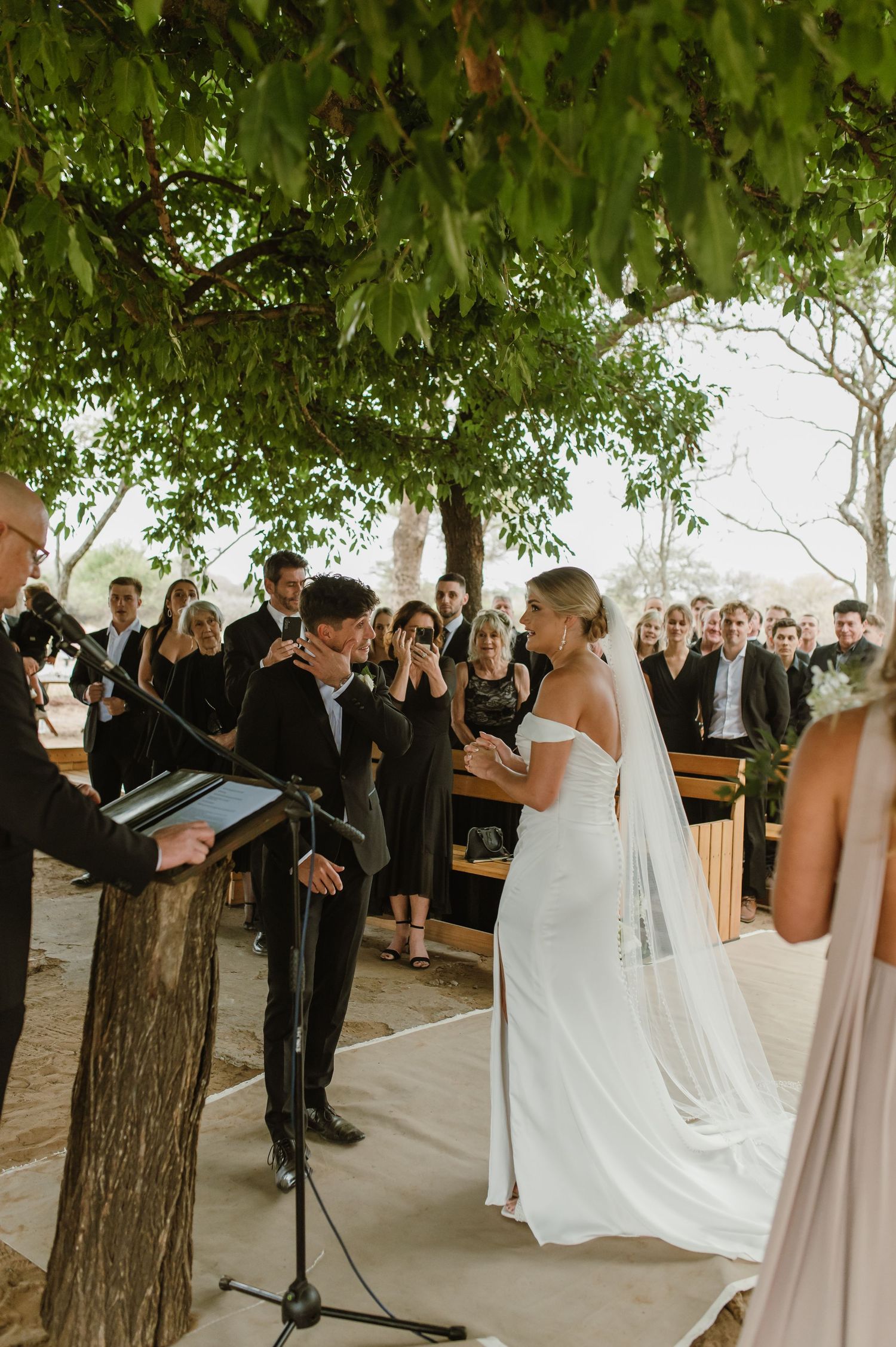 Wedding ceremony in progress under leafy green trees with guests watching.