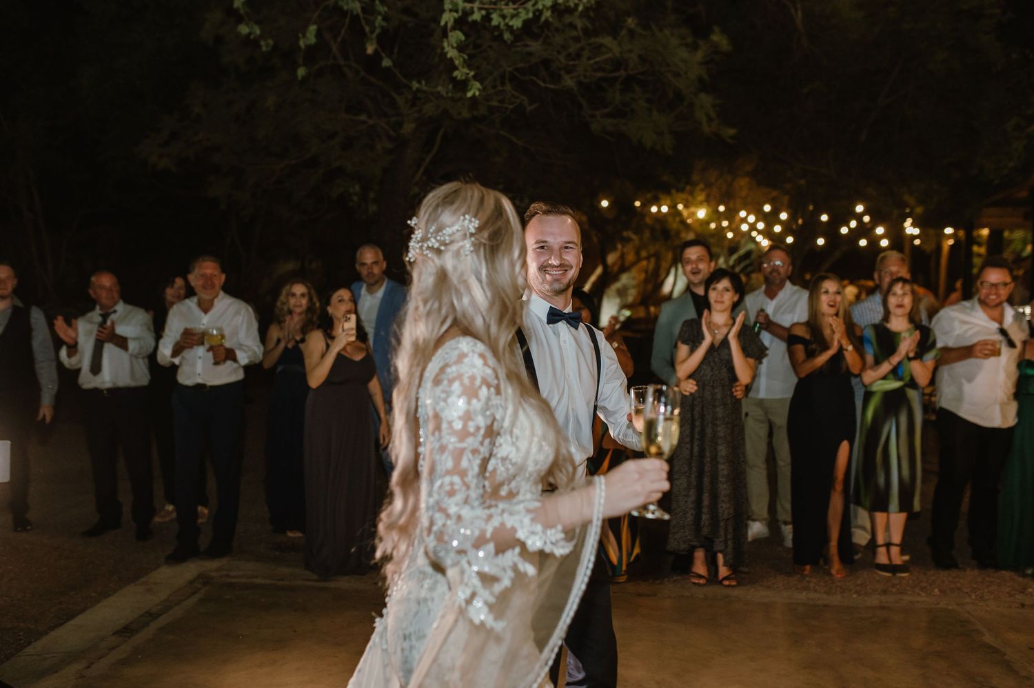 A couple shares their first dance at an outdoor evening wedding reception as guests watch and applaud.