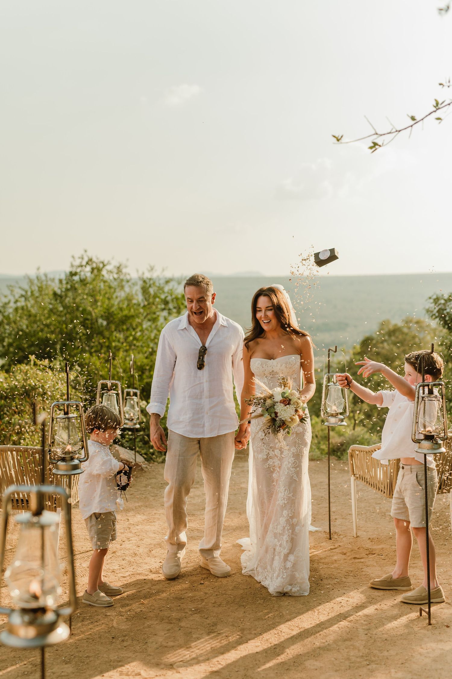 Bride and groom walk hand-in-hand with children at an outdoor wedding ceremony overlooking a scenic landscape.