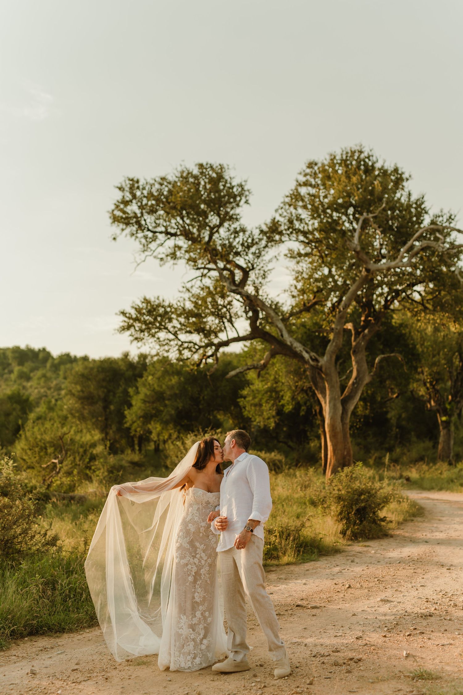 Bride and groom sharing a kiss on a sandy path surrounded by lush green trees during golden hour.