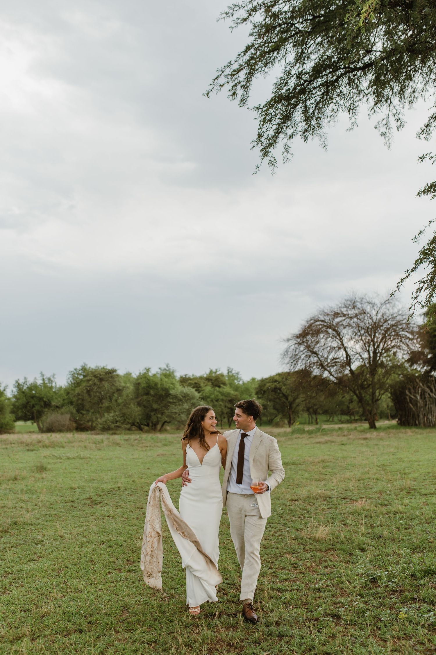 A wedding couple walks together in a grassy field wearing elegant white and beige attire.