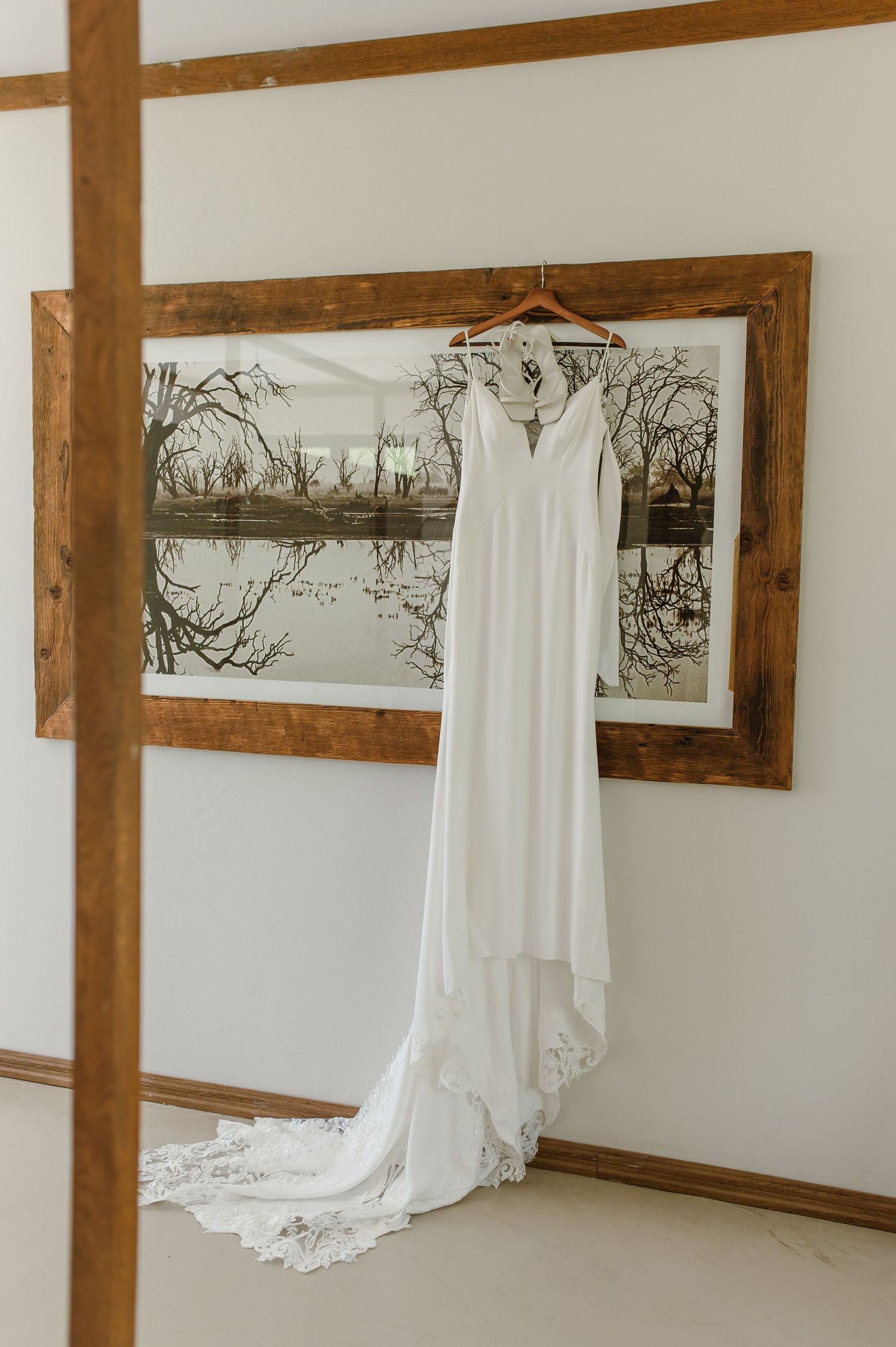 White wedding dresses hanging on wooden hangers against white wall with rustic framed artwork.