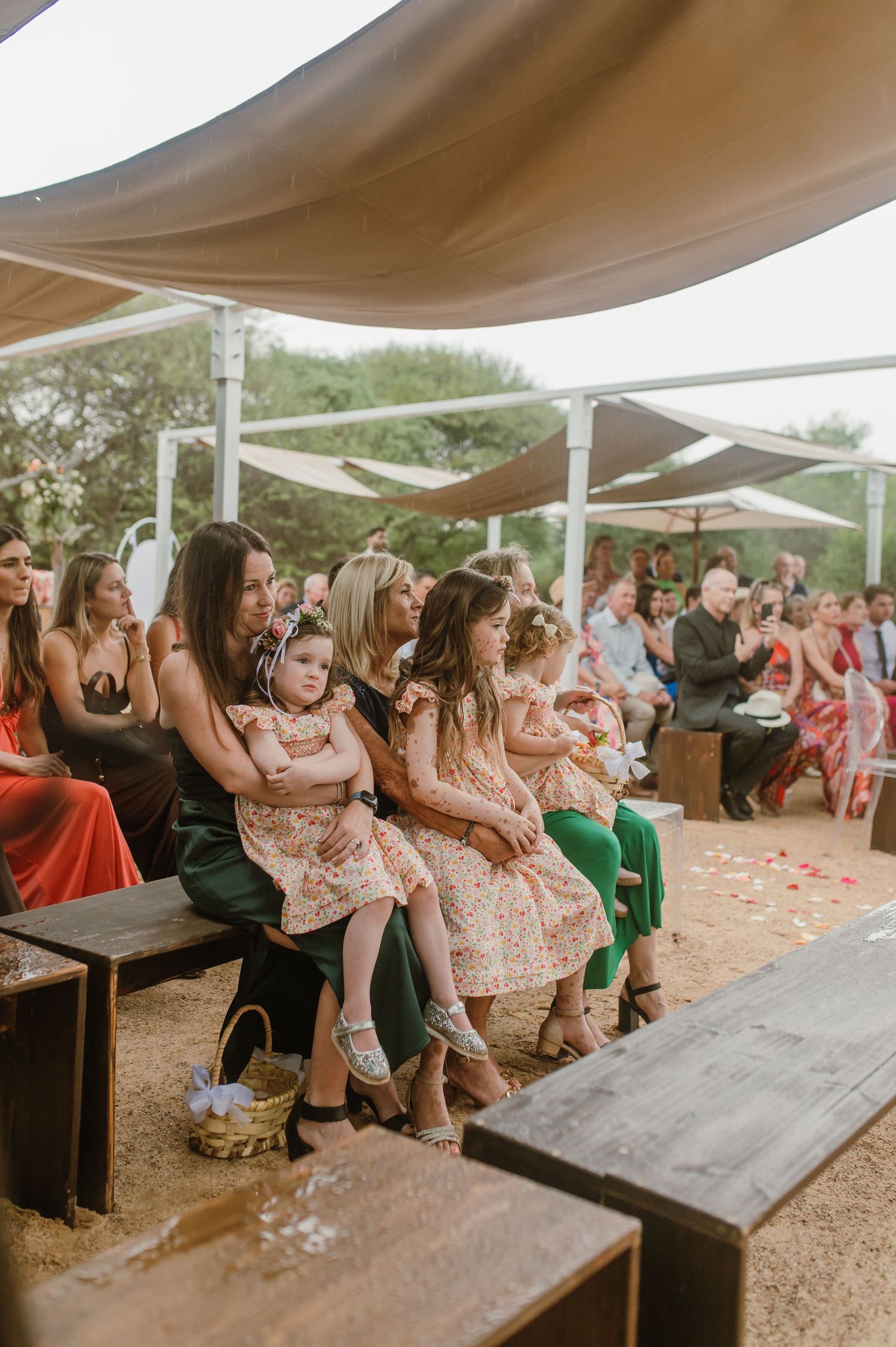 Wedding guests seated on wooden benches under a canopy at an outdoor desert venue.