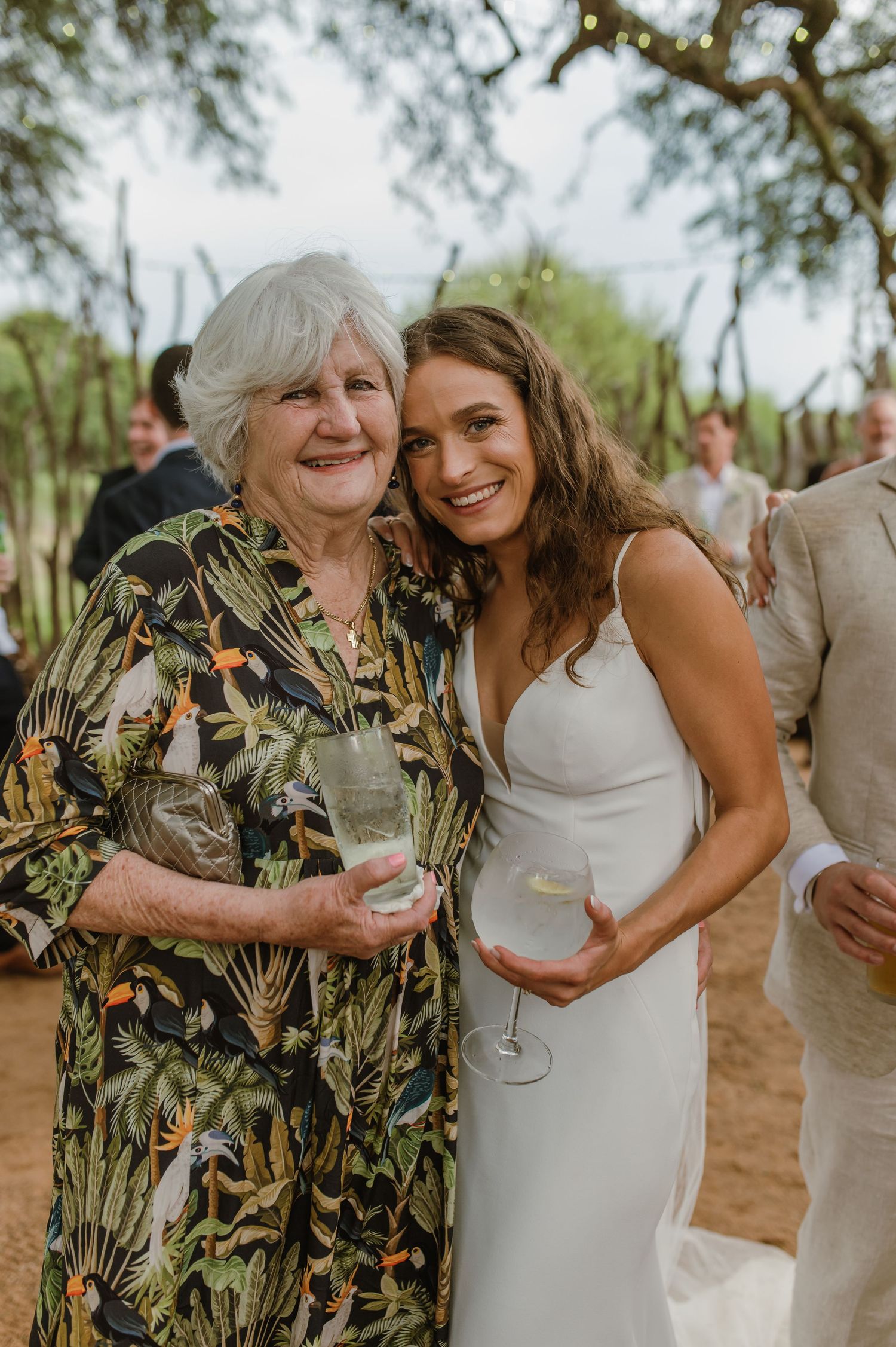Wedding guests share joyful moments and drinks under trees at an outdoor celebration.