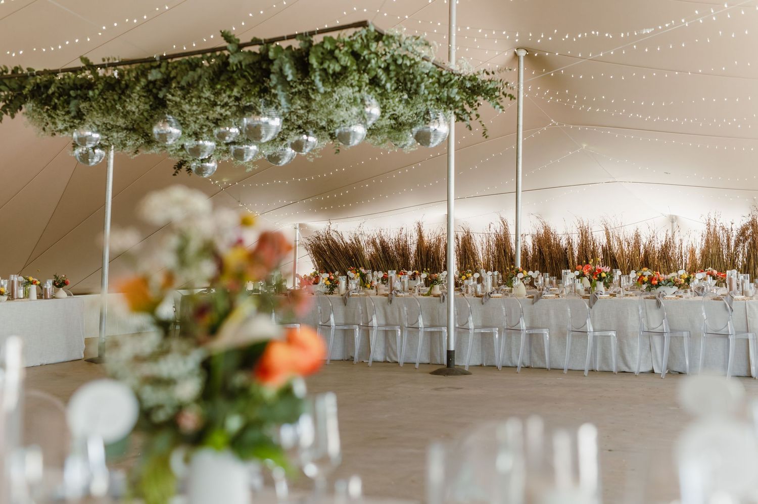 Wedding reception tent interior with string lights and floral decorations hanging from ceiling.