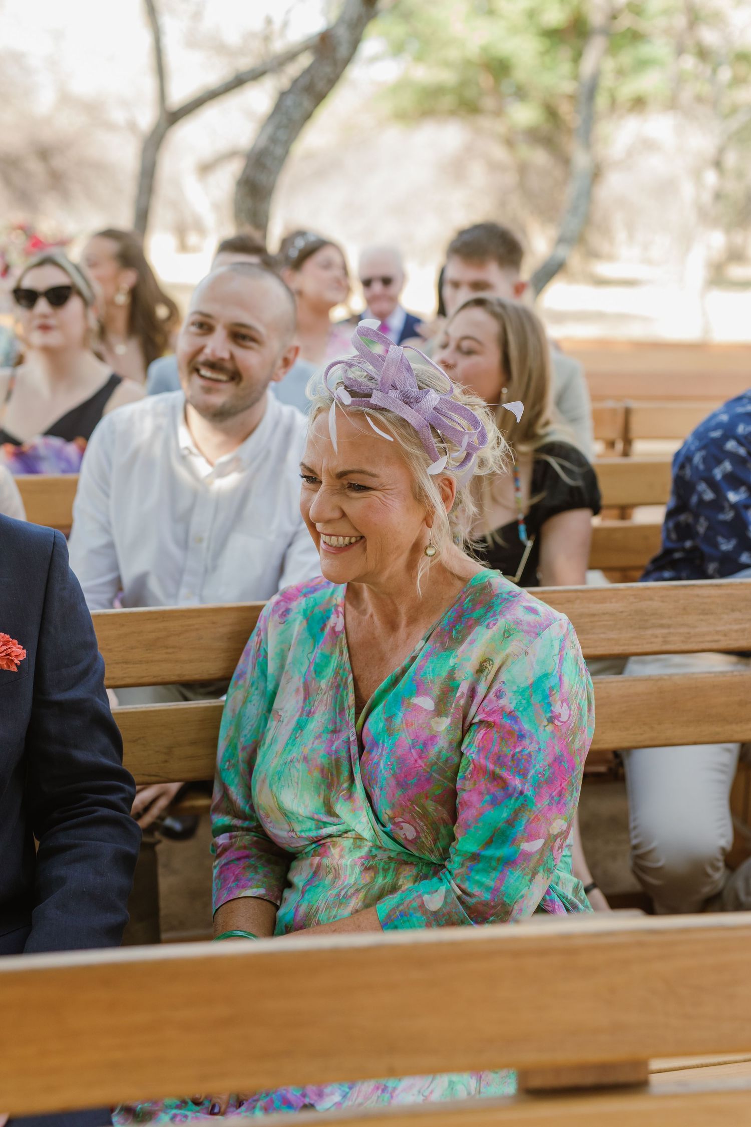 Wedding guests in colorful attire share laughs while seated on wooden benches.