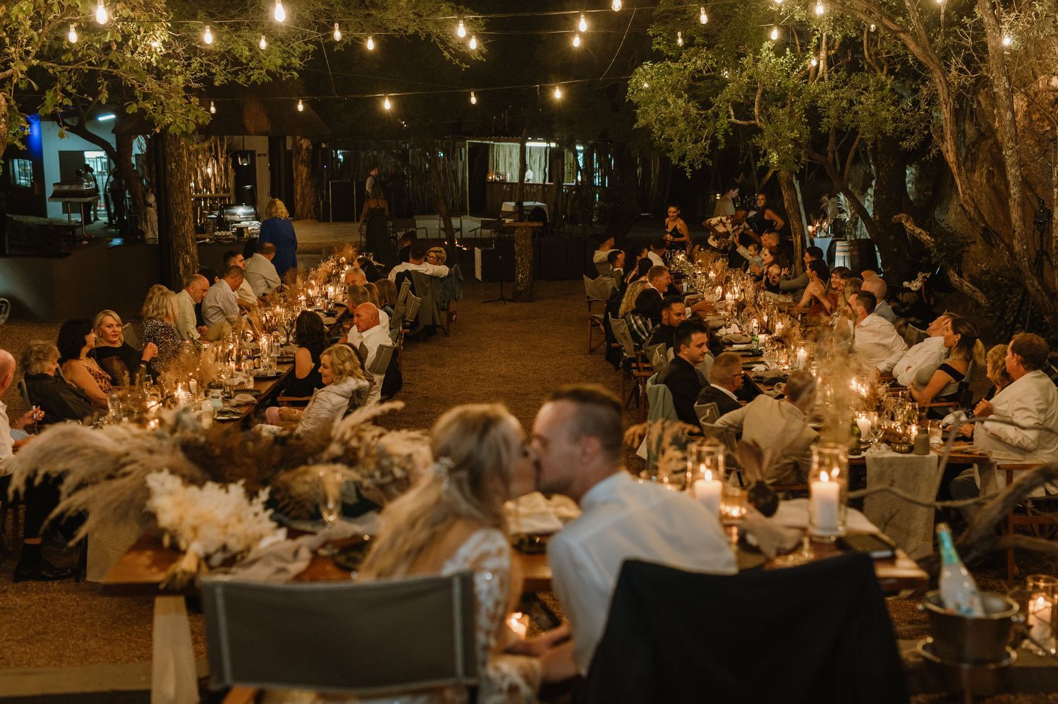 Evening outdoor wedding reception with guests seated at long tables under string lights.