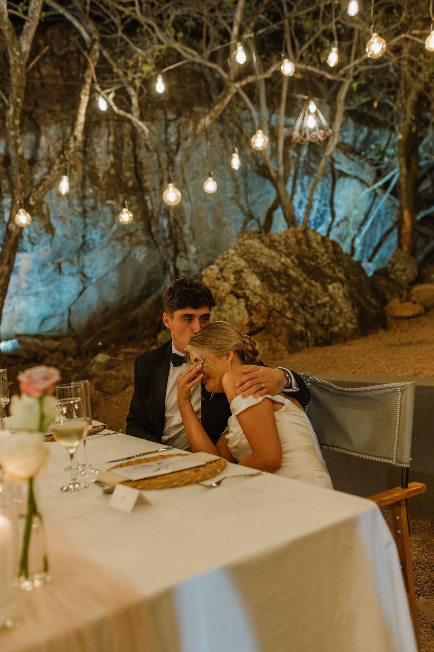Wedding reception dinner table decorated with white linens under twinkling lights in nature.