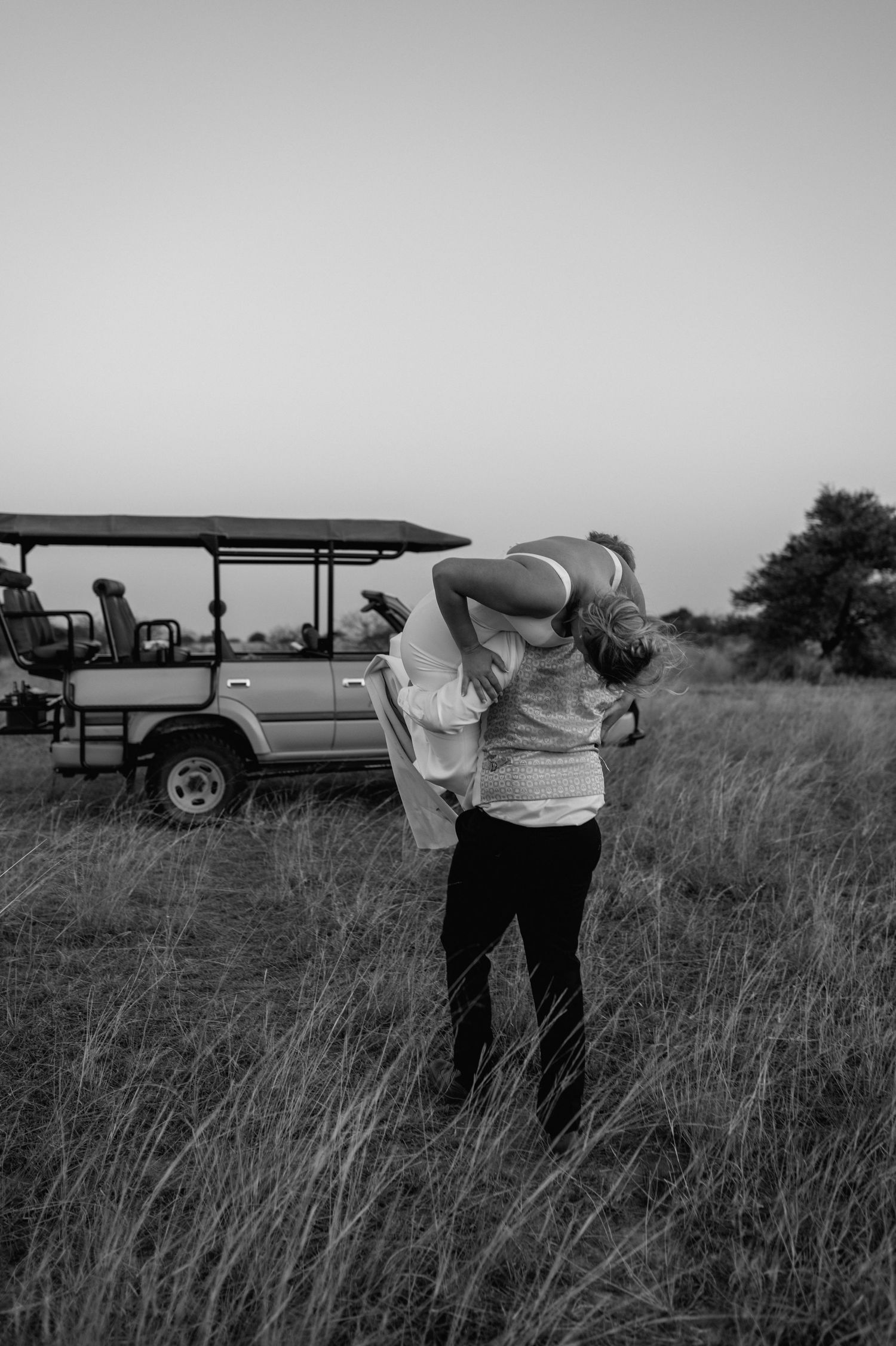 A black and white photo of a couple standing near a safari vehicle in tall grass.