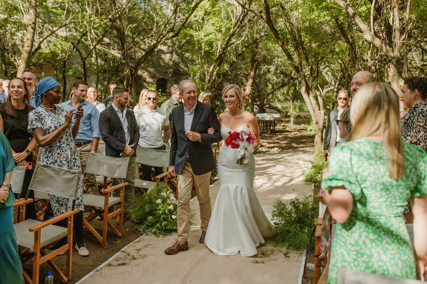 Guests watch a wedding ceremony on a sunlit garden path surrounded by lush greenery.