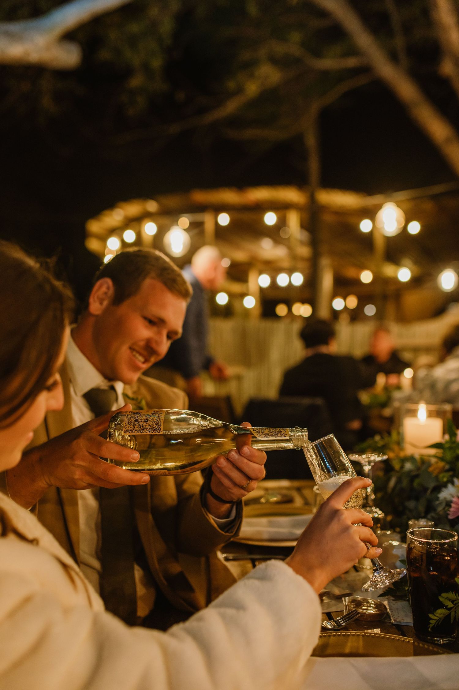Wedding guests raise glasses for a toast during an evening reception under warm lighting.