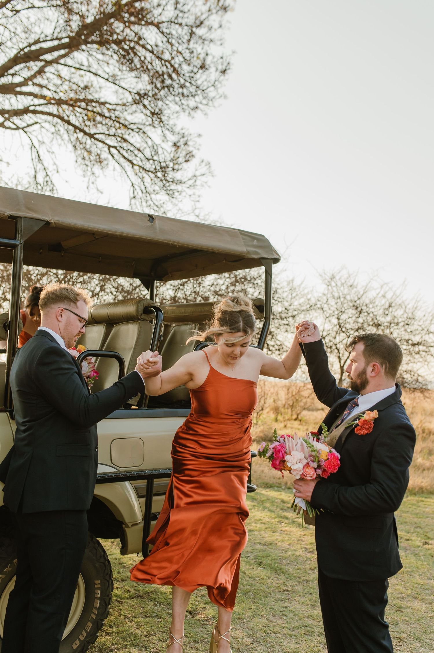 Bridal party in burnt orange dresses dances and laughs together near a vintage vehicle.