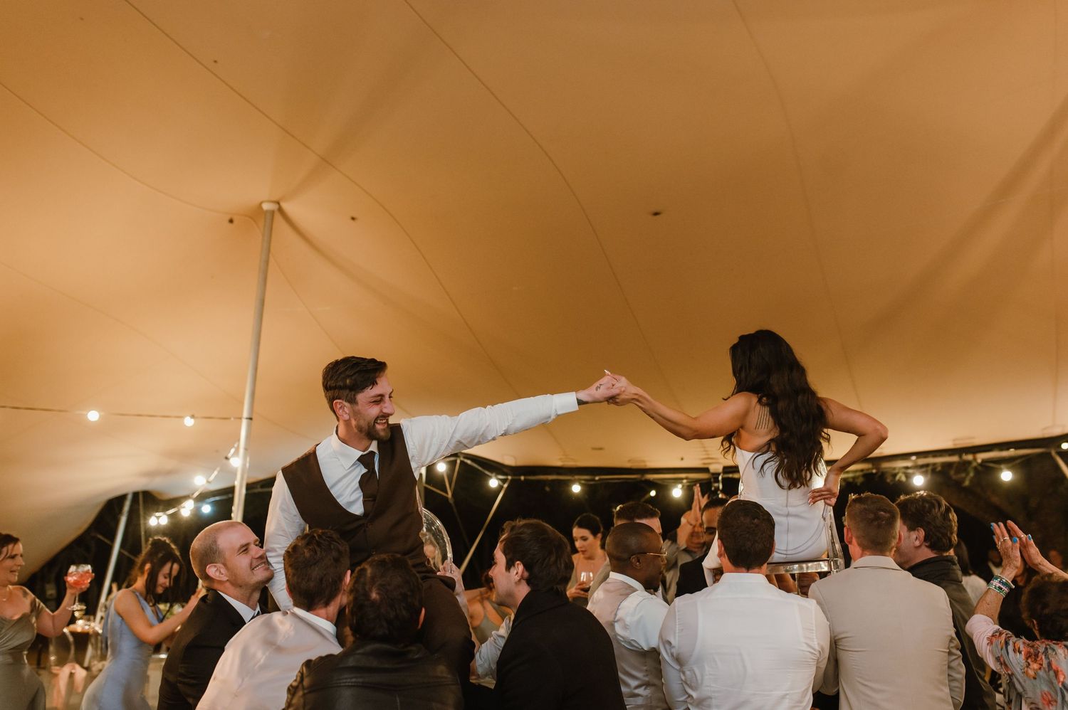 Two people hold hands while being lifted by guests during a festive wedding celebration under a tent.