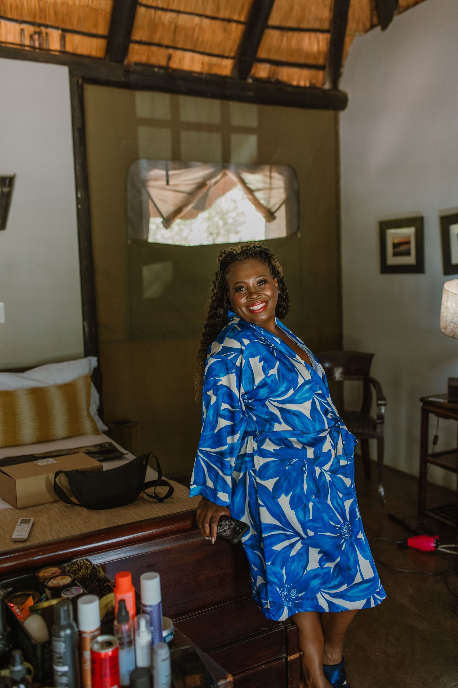 Woman in vibrant blue floral print outfit sitting in rustic room with thatched roof.