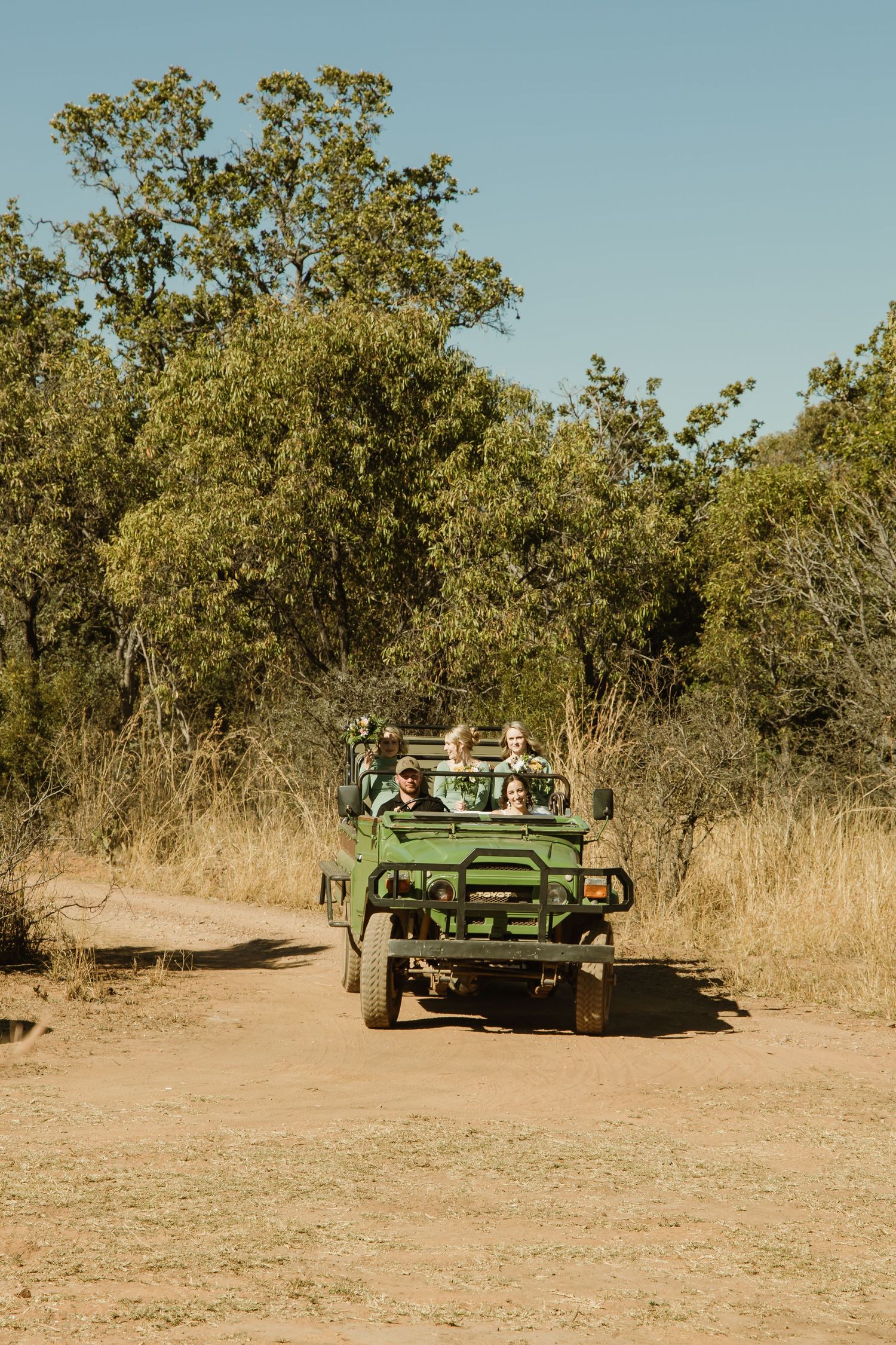 Green safari vehicle parked on a dirt path surrounded by dry brush and trees in a natural setting.