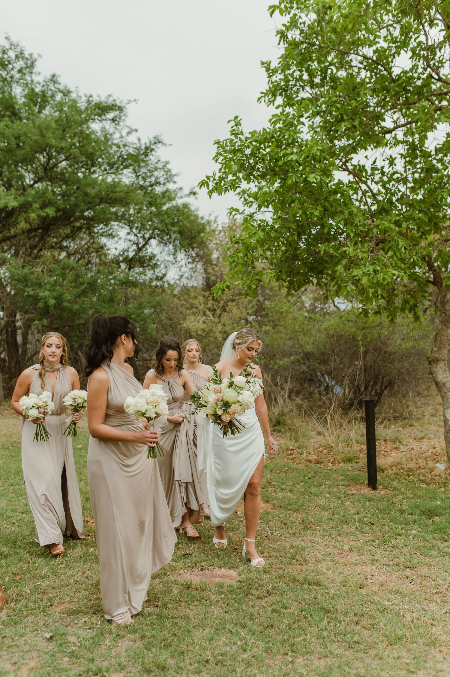 Bridal party in neutral dresses walks together through natural outdoor setting carrying bouquets.
