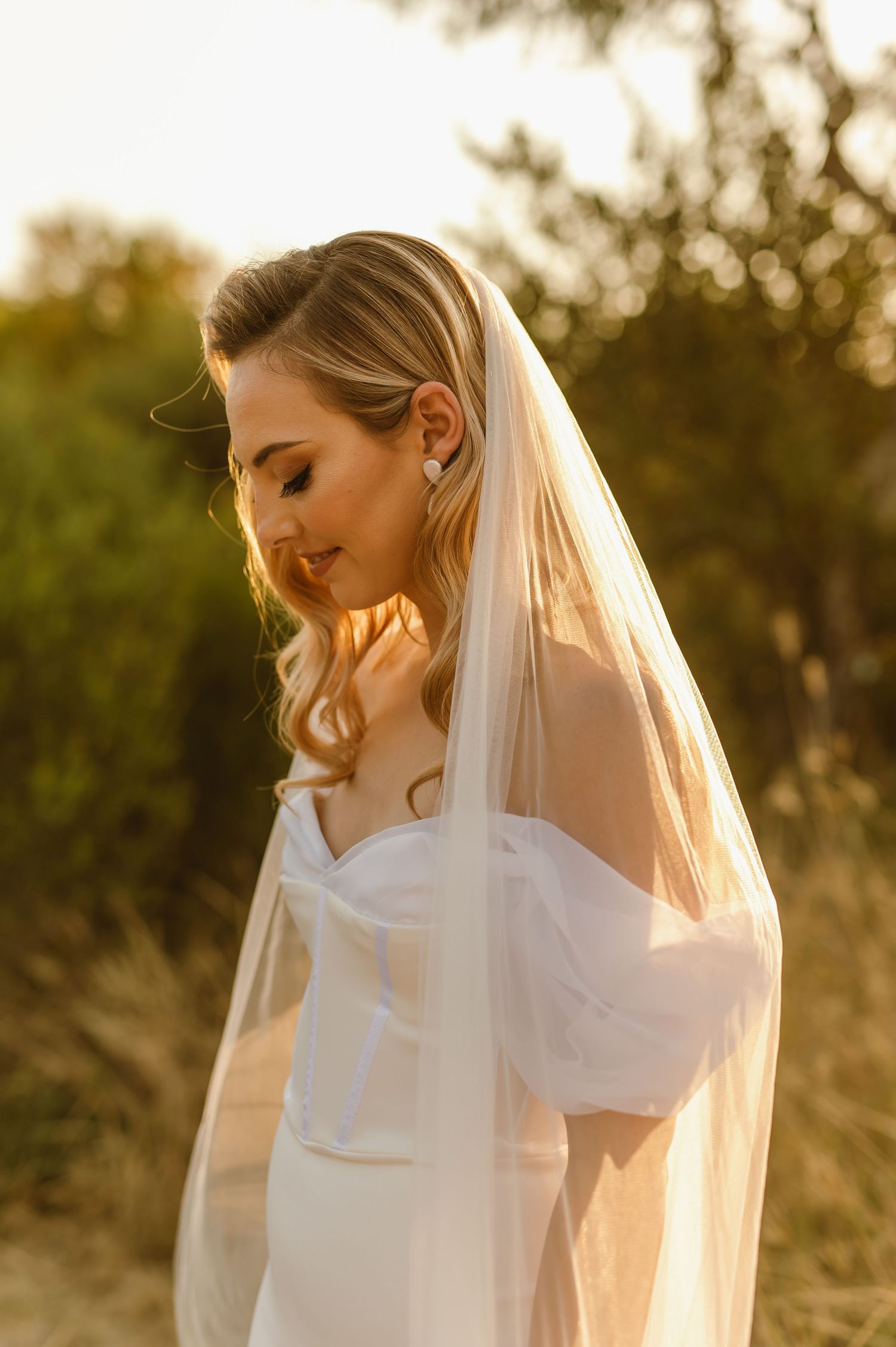 A bride in a white dress poses during golden hour sunset with her veil flowing in the breeze.