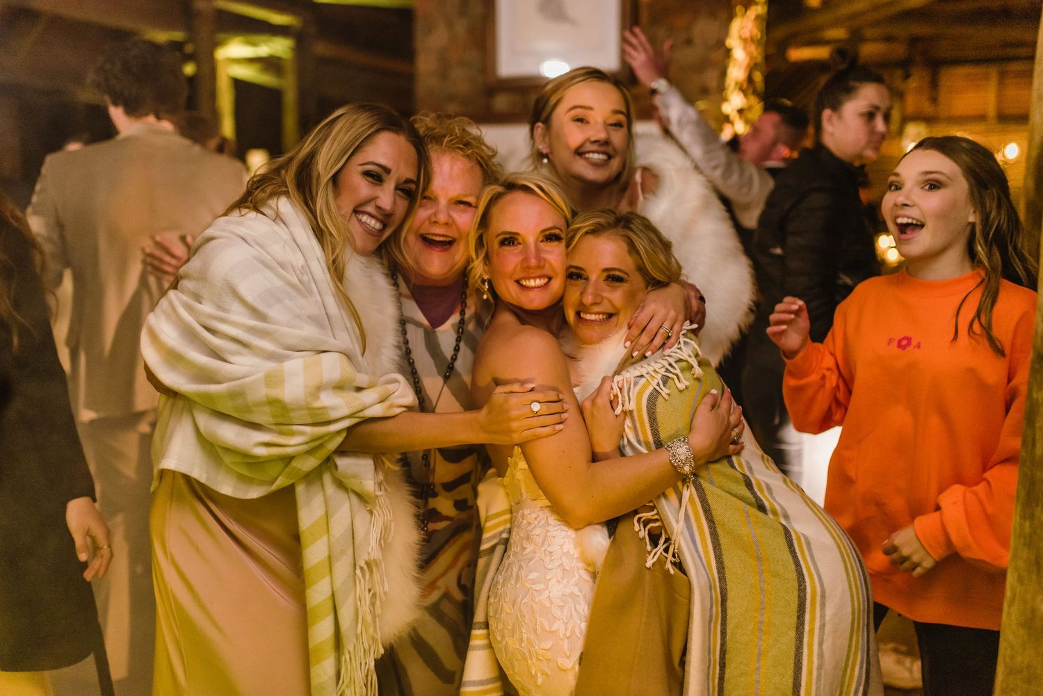 Wedding guests embrace and celebrate during a lively barn reception under string lights.