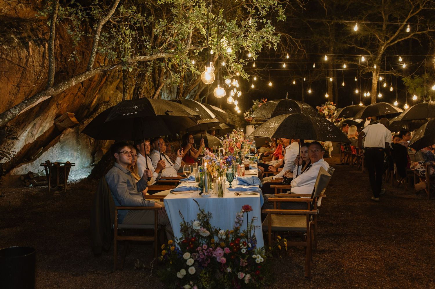 Wedding guests dine under string lights at outdoor reception, some holding umbrellas in the warm evening.