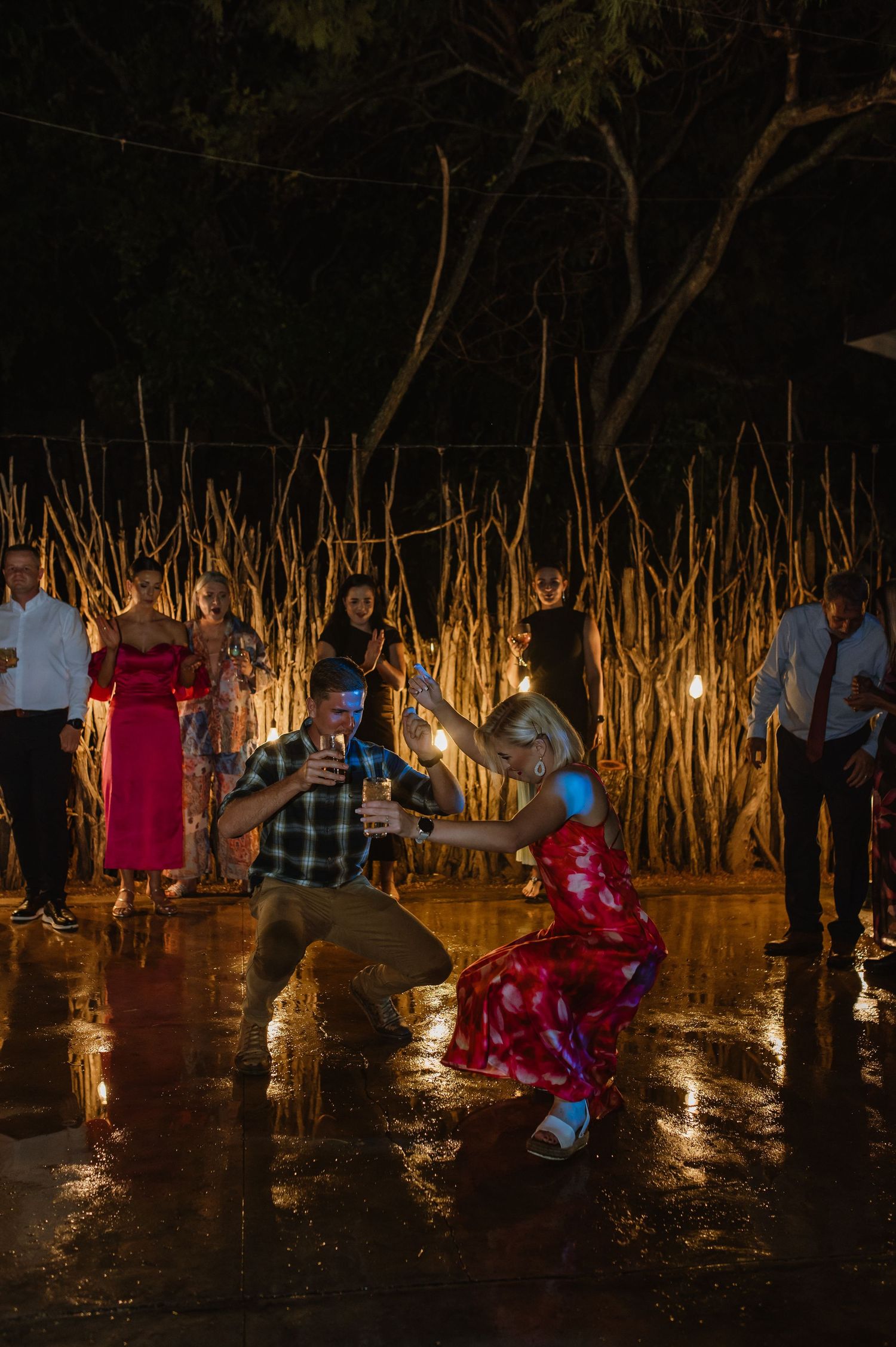Guests dance energetically on rain-soaked outdoor wedding dance floor at night under warm string lights.
