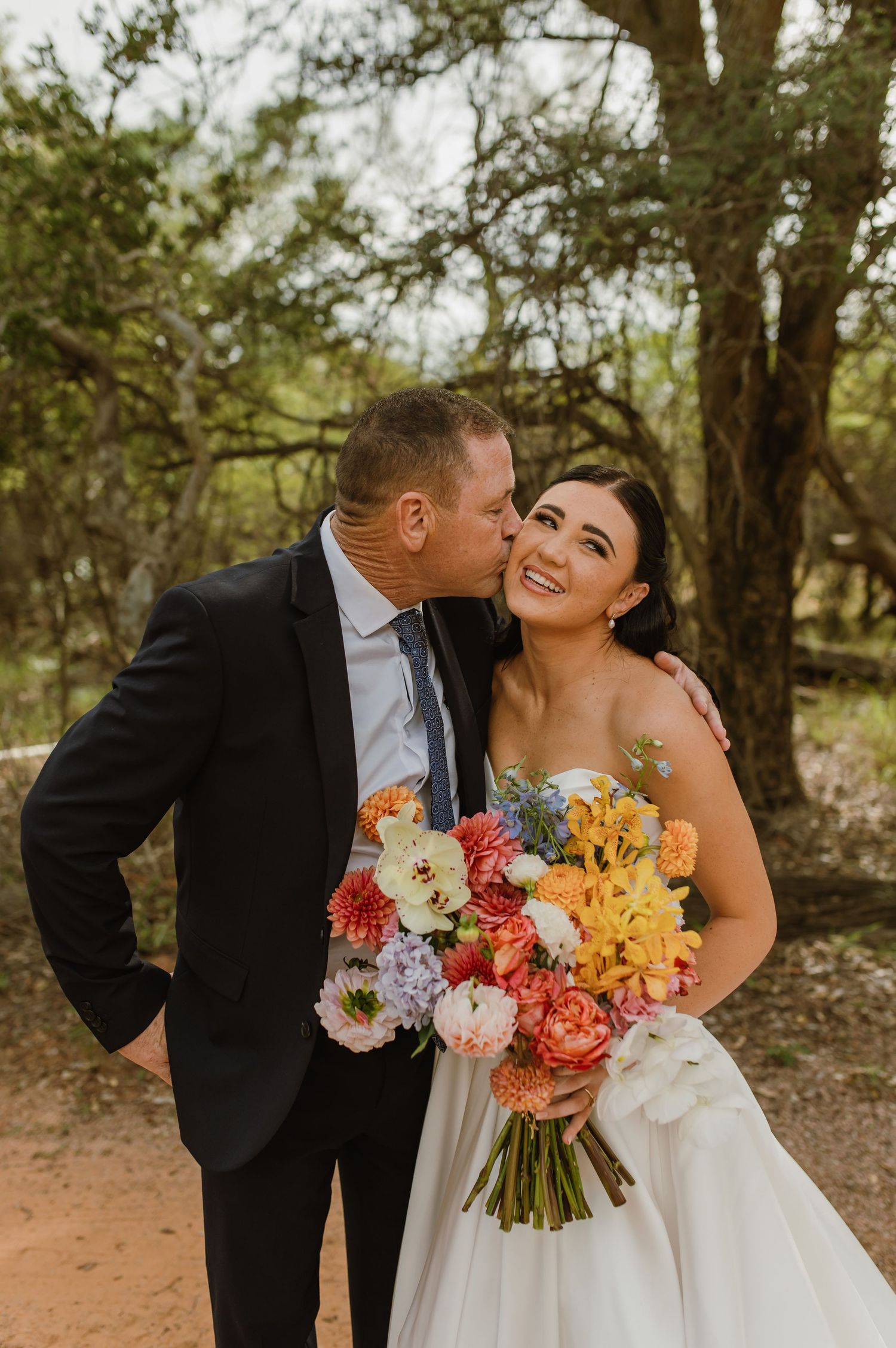 Groom kissing smiling bride on cheek as she holds colorful wedding bouquet outdoors.
