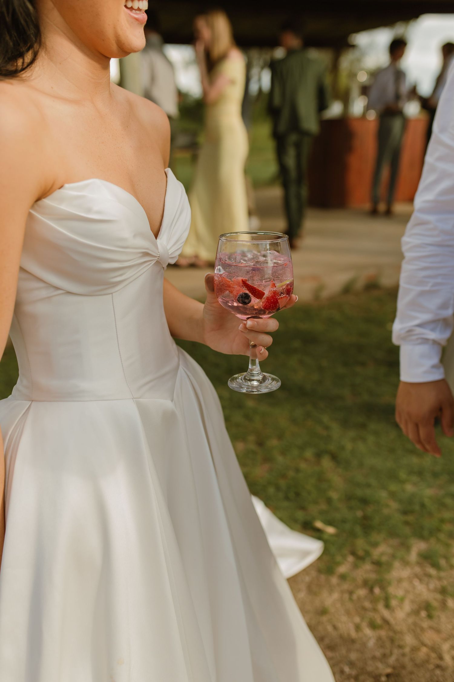 Bride in strapless white gown holding a berry-filled cocktail glass at an outdoor wedding reception.