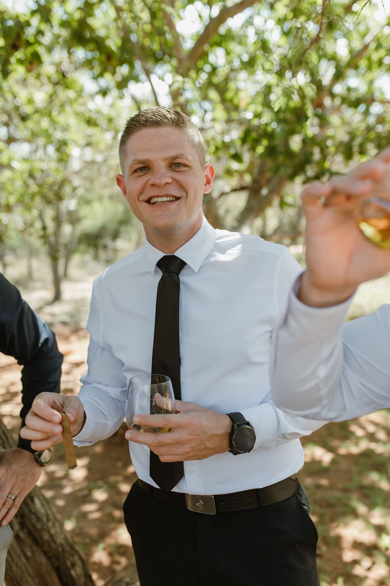 Smiling groom in white shirt and black tie raises a glass during outdoor wedding celebration.