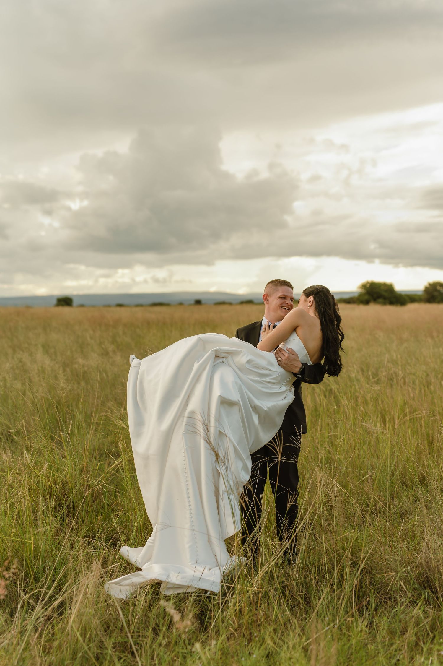 Groom lifts bride in white gown in golden grass field under dramatic cloudy sky at sunset.