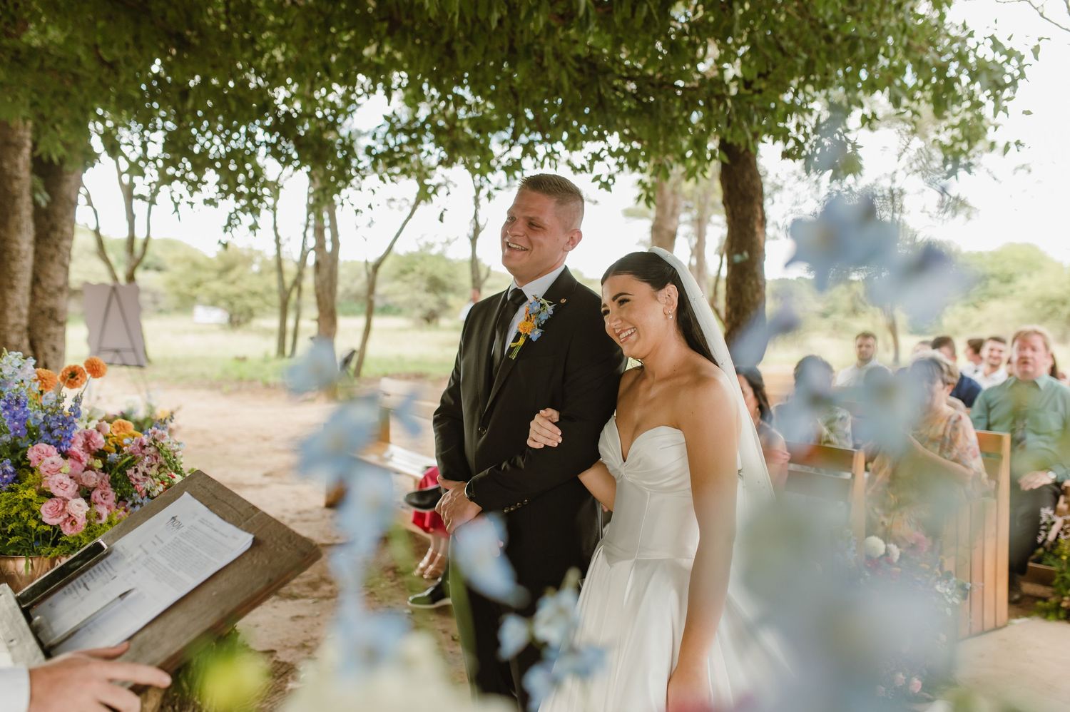 Bride and groom standing at outdoor wedding ceremony beneath a lush green tree.