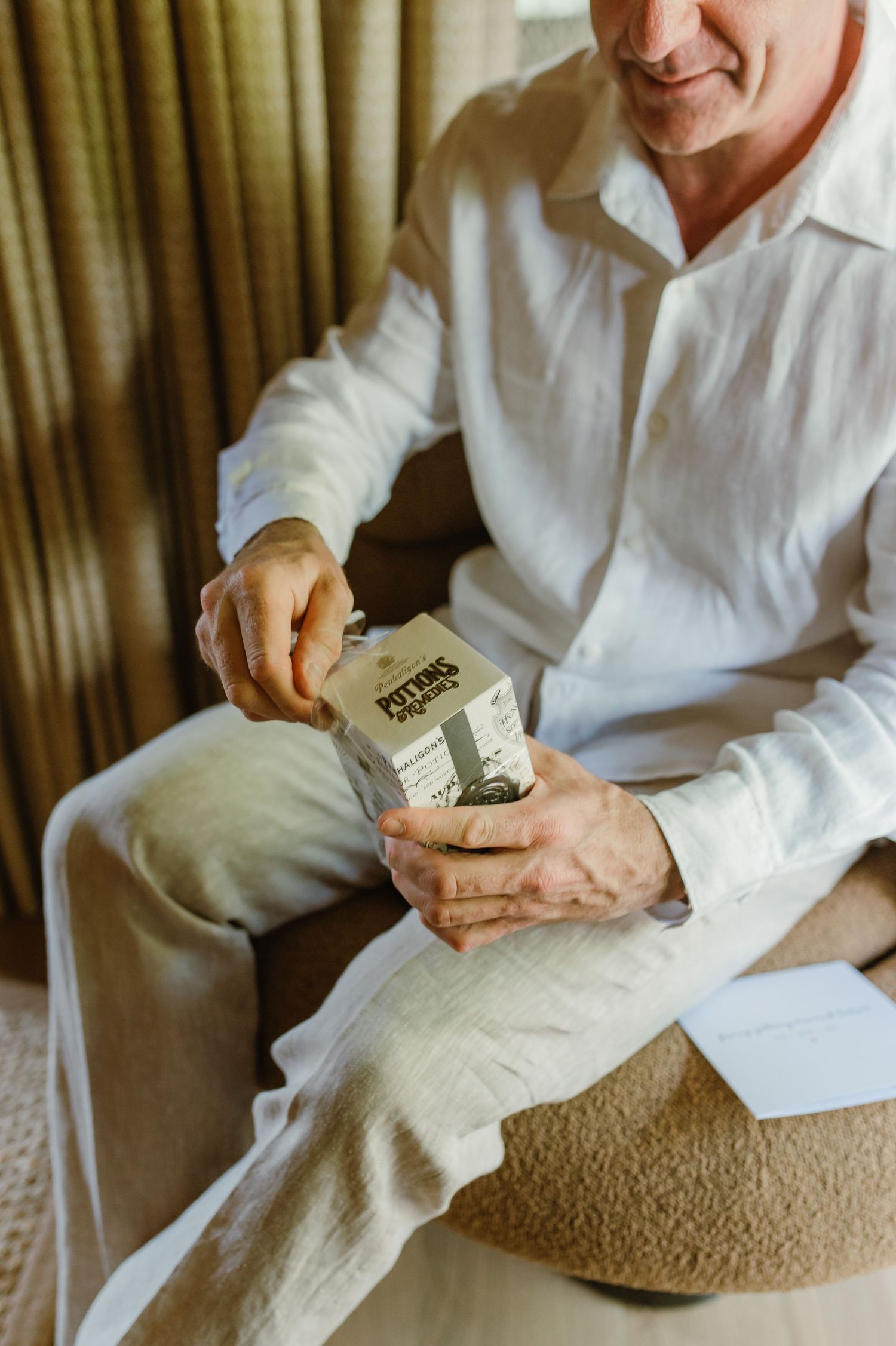 Man in white linen shirt sitting in chair opening a bottle of Pittres cologne fragrance.