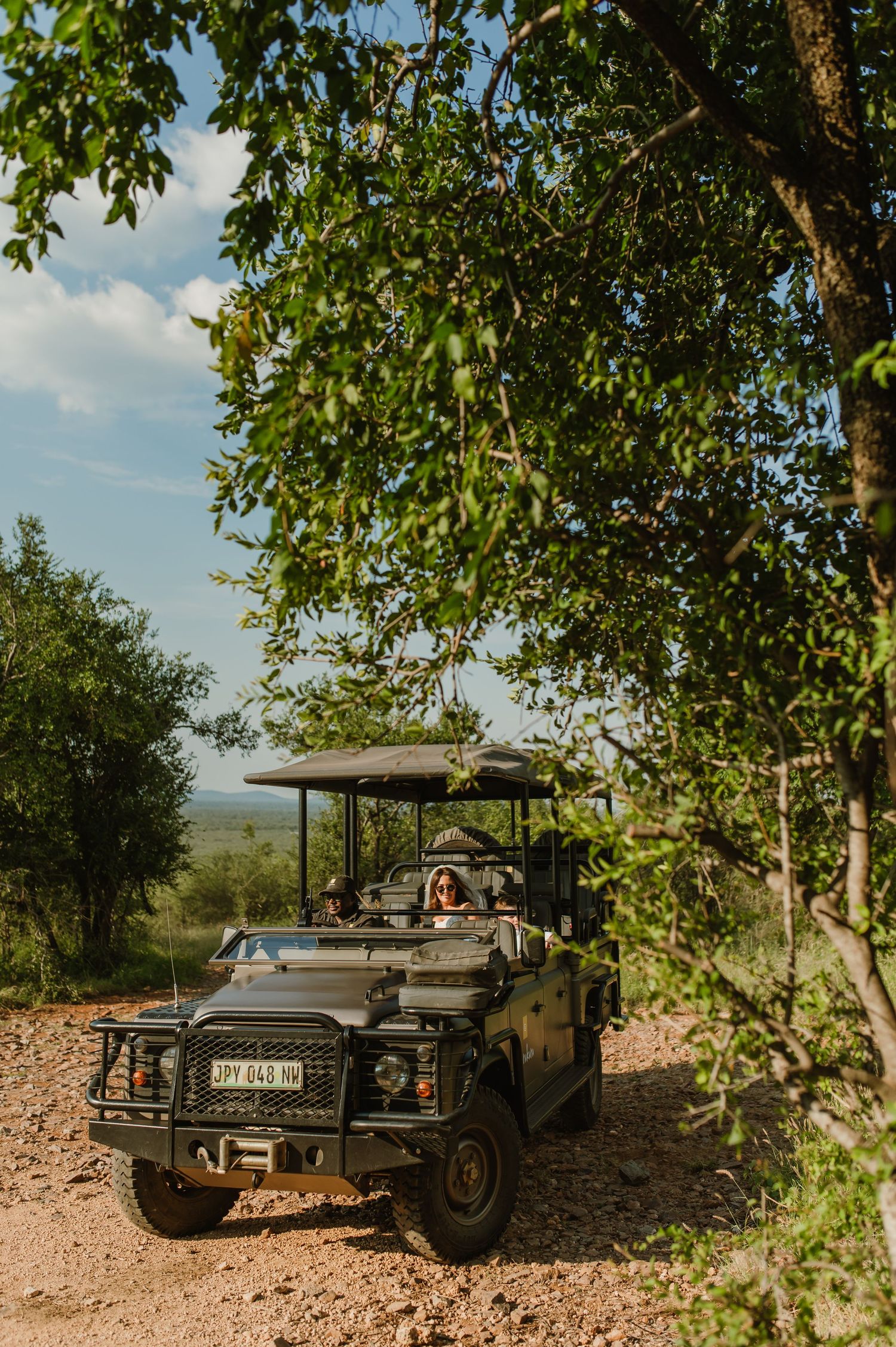 Bride riding in open-top safari jeep through lush green trees on dirt path.