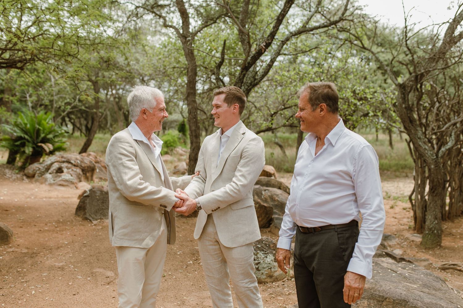 Three men in business attire having a conversation while standing on a dirt path surrounded by trees in a natural setting.