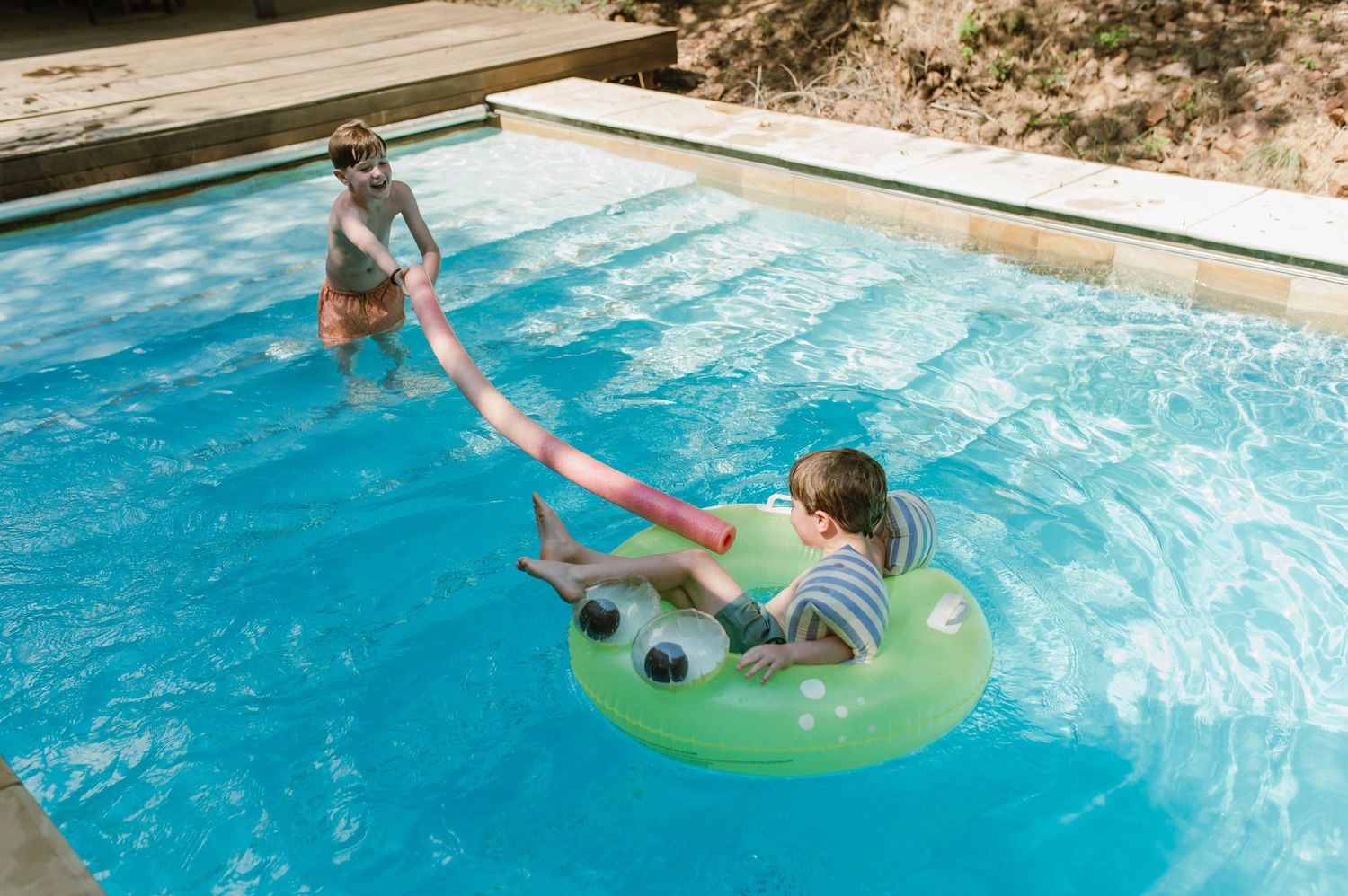 Two children playing in a outdoor swimming pool, one on a green inflatable ring being pulled.