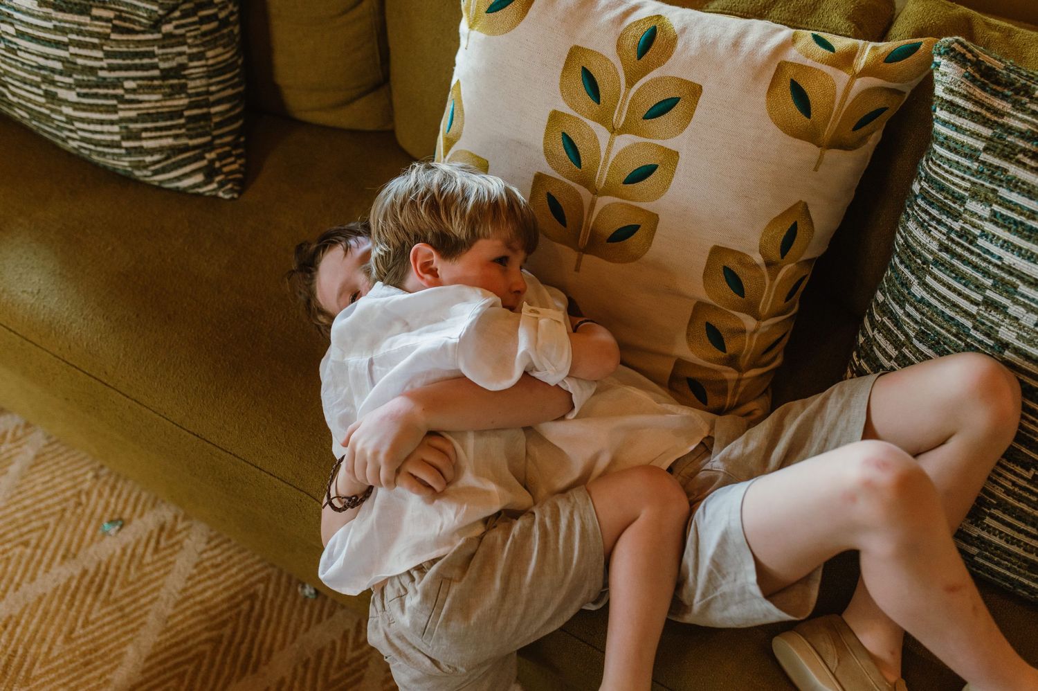 Two young boys cuddle together on a yellow sofa with decorative leaf-print pillows.