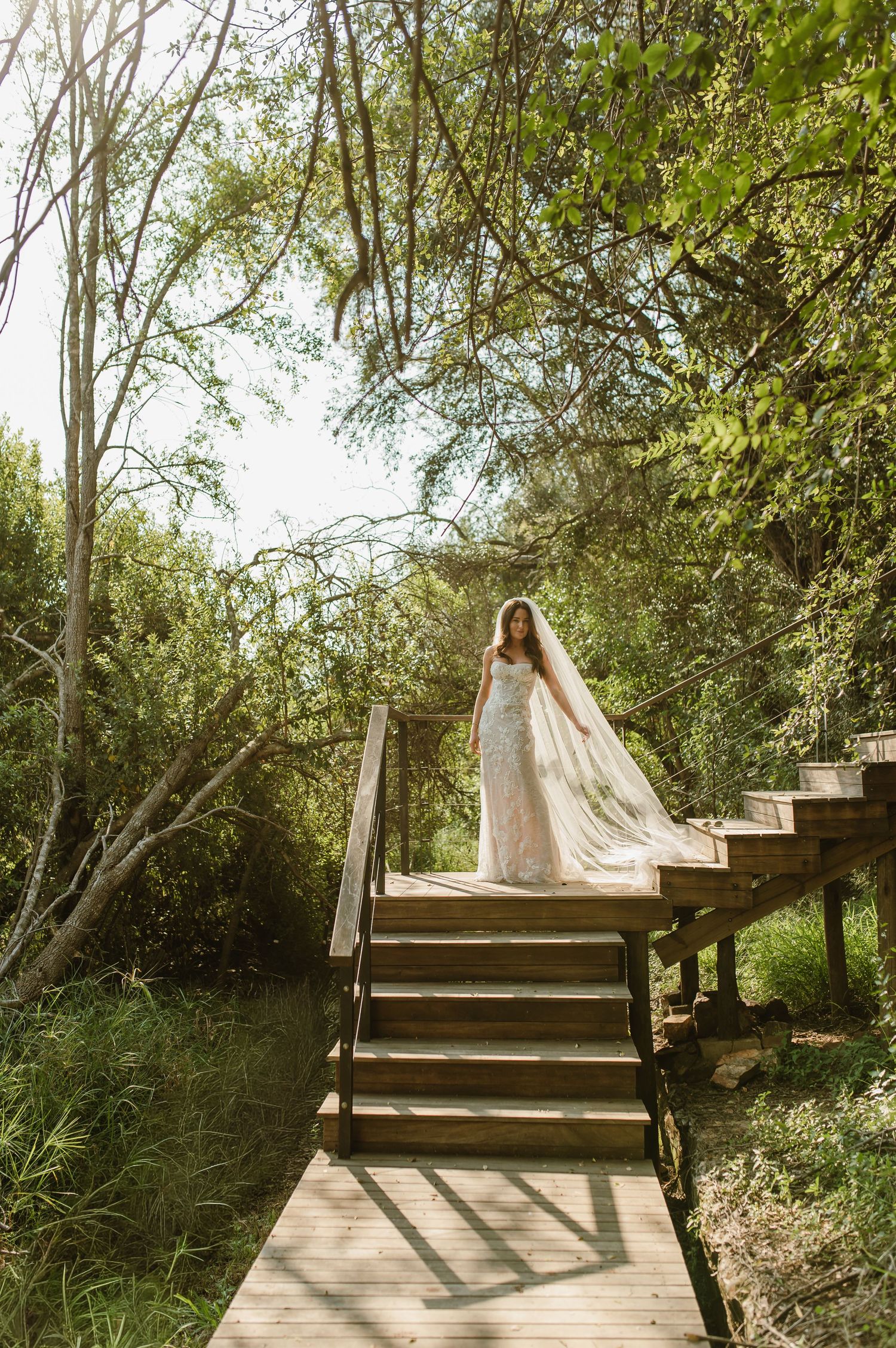 Bride in strapless lace gown with flowing veil standing atop wooden staircase surrounded by lush green trees.
