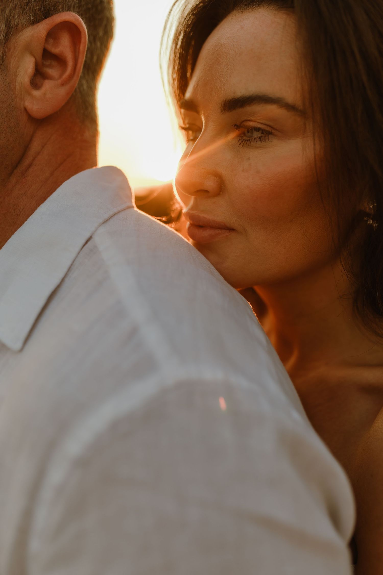 Close-up of bride resting her head on groom's shoulder bathed in warm golden sunset light.