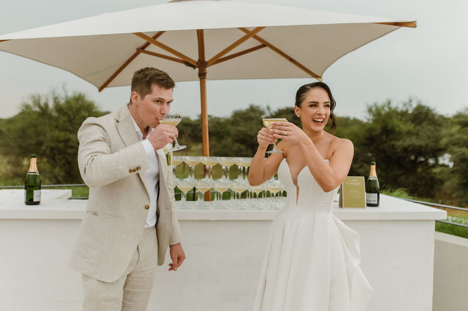 A couple shares a celebratory moment while drinking champagne under a white umbrella on their wedding day.
