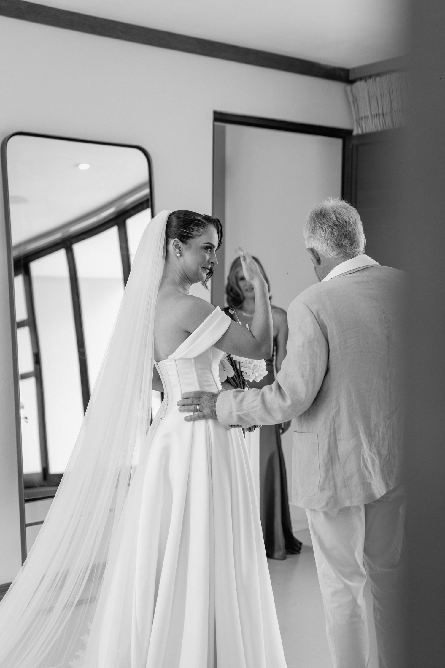 Black and white photograph showing final wedding dress adjustments in a sunlit room.