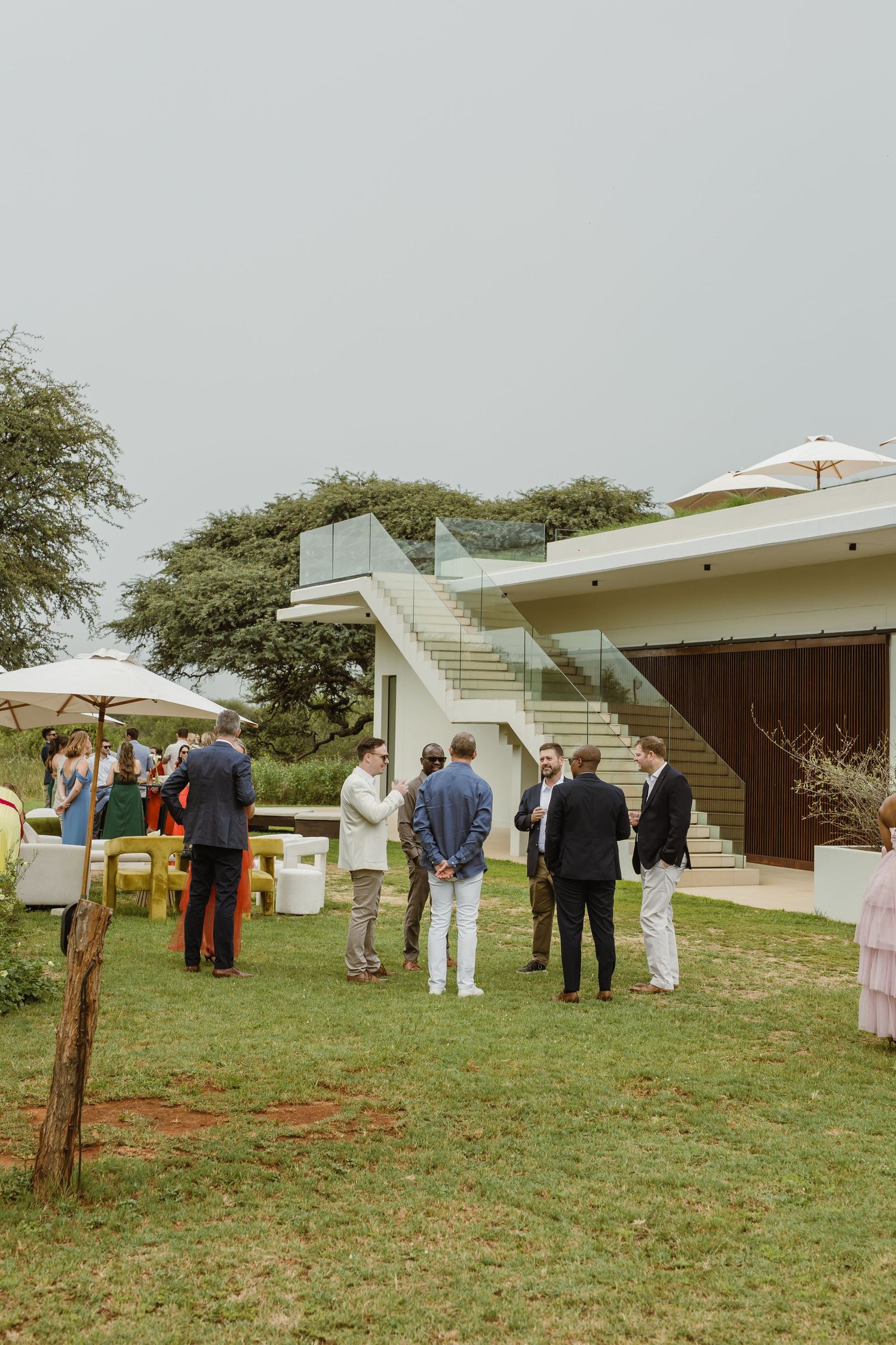 Wedding guests gather outside a modern white building on a grassy lawn during an outdoor ceremony.