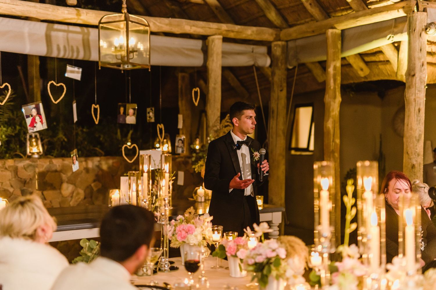Wedding toast being given in a rustic barn venue decorated with hanging lights and candles.