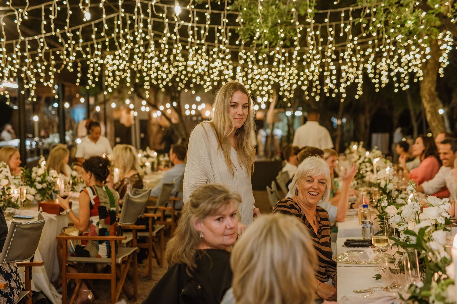 Wedding reception guests seated at long tables under twinkling fairy lights in an enchanted outdoor setting.