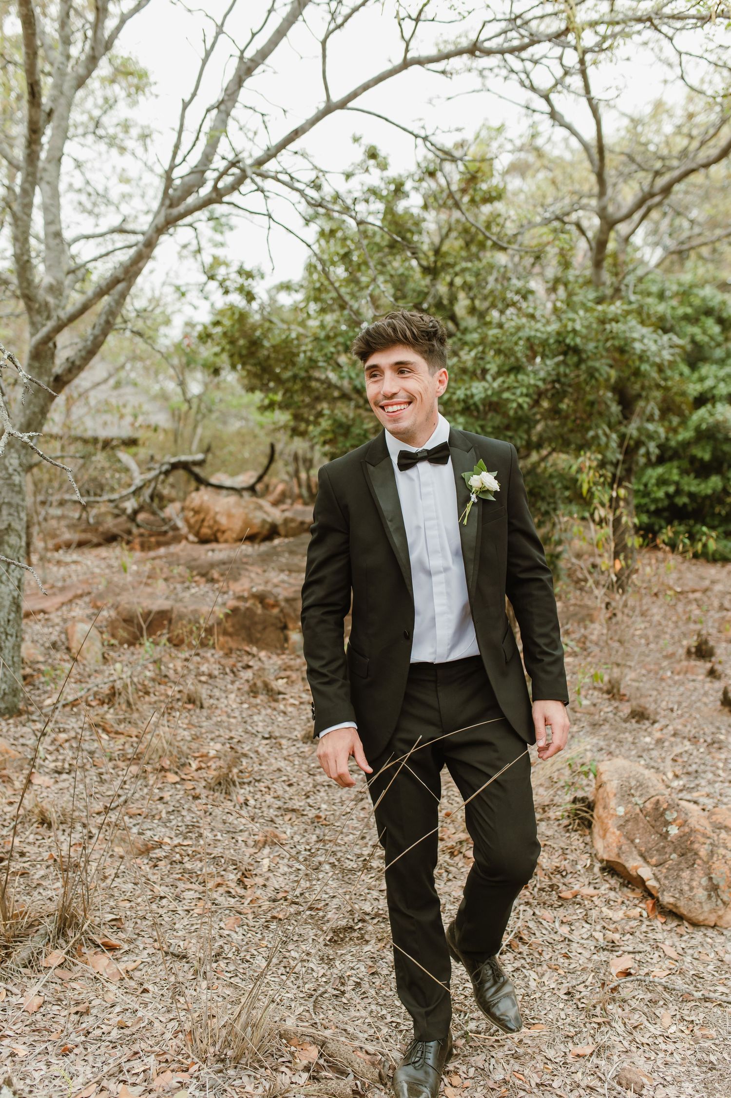 Groomsmen in black tuxedos pose casually on a rocky outdoor trail surrounded by trees and foliage.