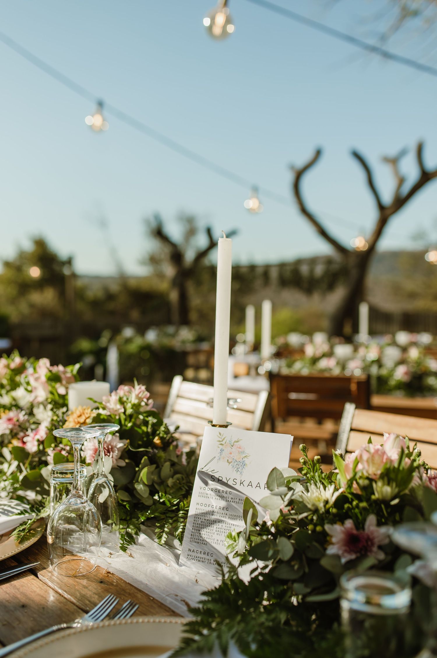 Close-up of wedding table setting with white candles, greenery, and floral arrangements under string lights.